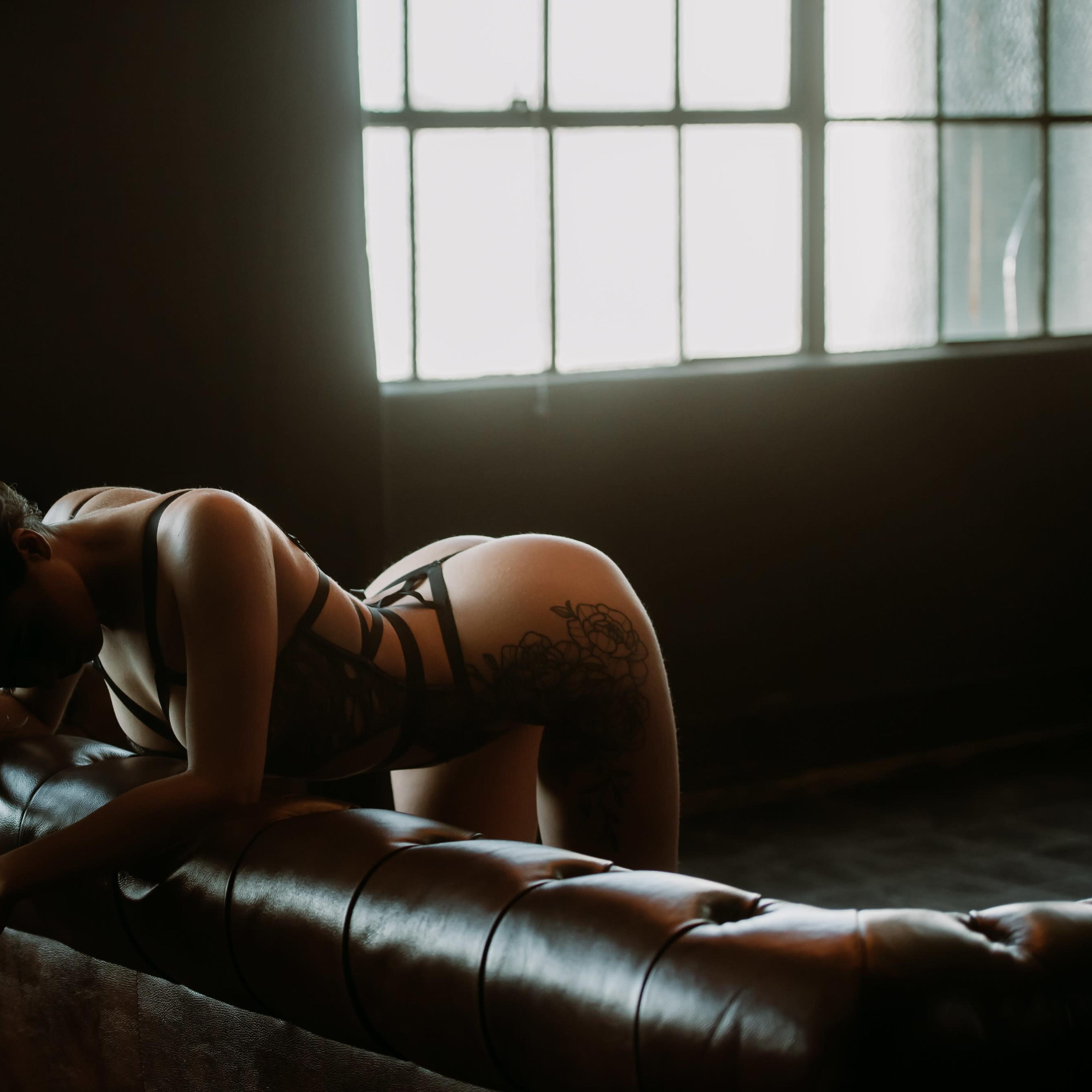 A person in lingerie leans over a leather couch in a dimly lit room near a large window.