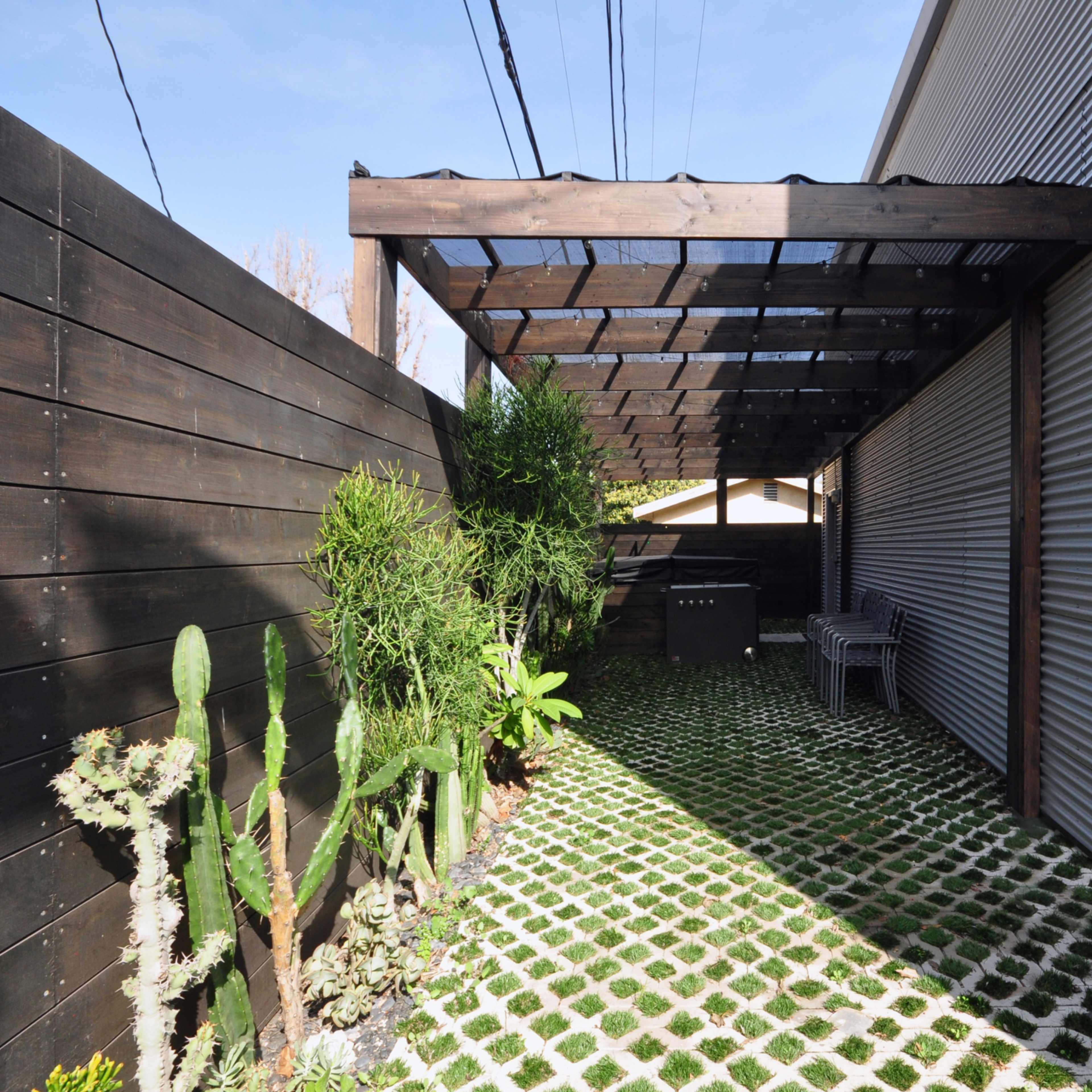 A narrow outdoor pathway features a gravel-like surface with green vegetation, shaded by a wooden pergola and flanked by a metal wall.