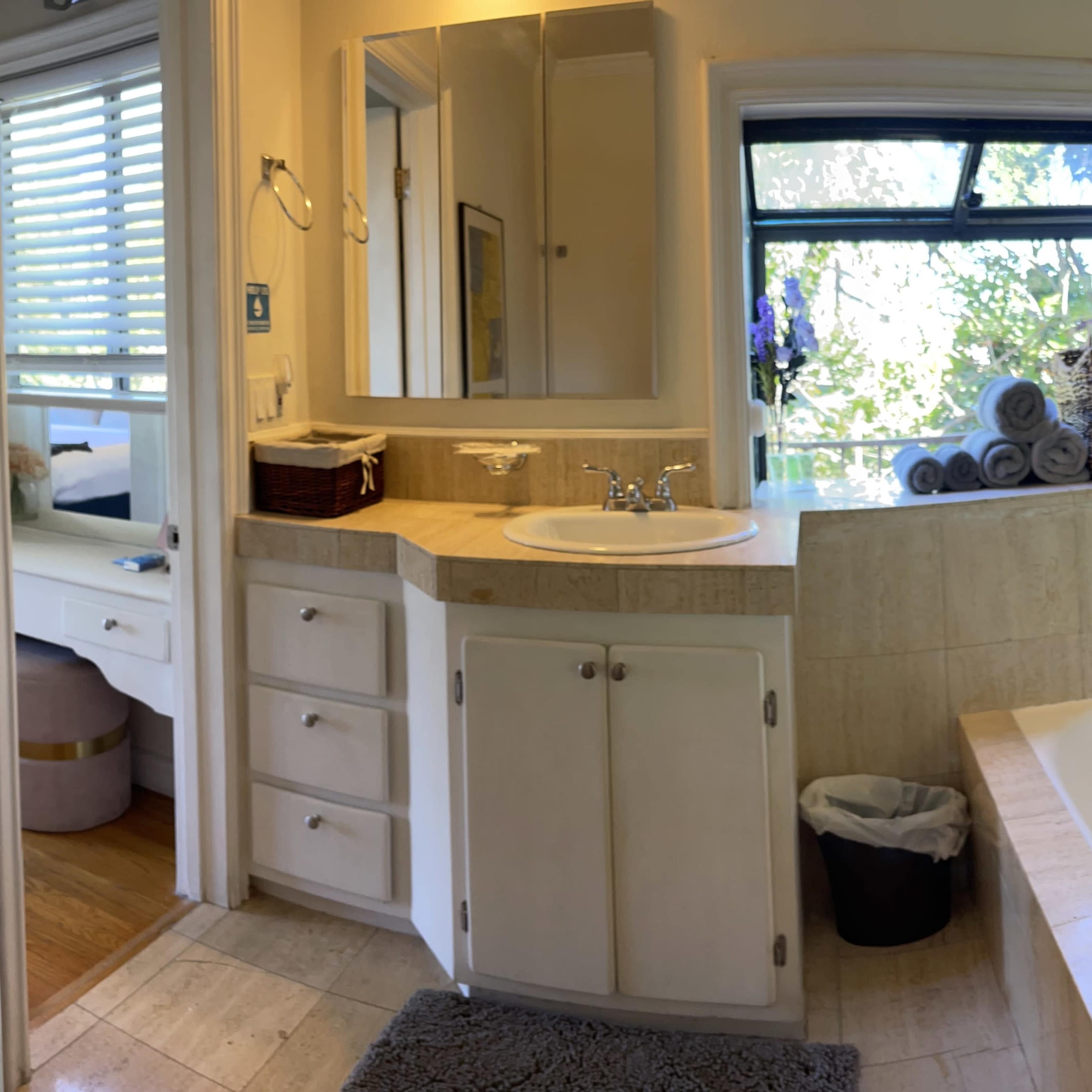 The bathroom features a sink area with a large mirror, light-colored cabinets, and a window allowing natural light, alongside a bathtub and neatly organized towels.