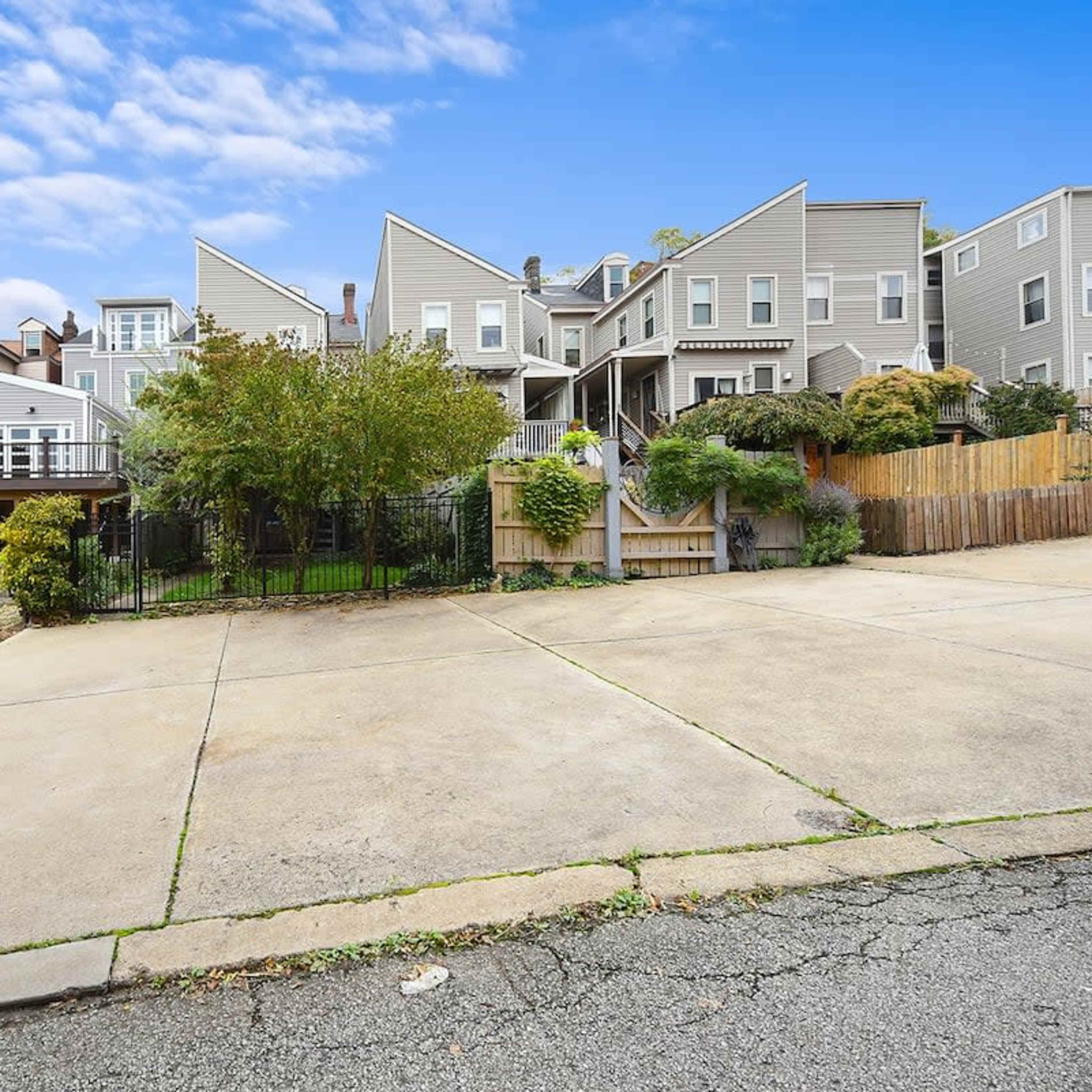 A paved area in front of multiple residential buildings with pitched roofs and a landscaped yard behind a black fence.
