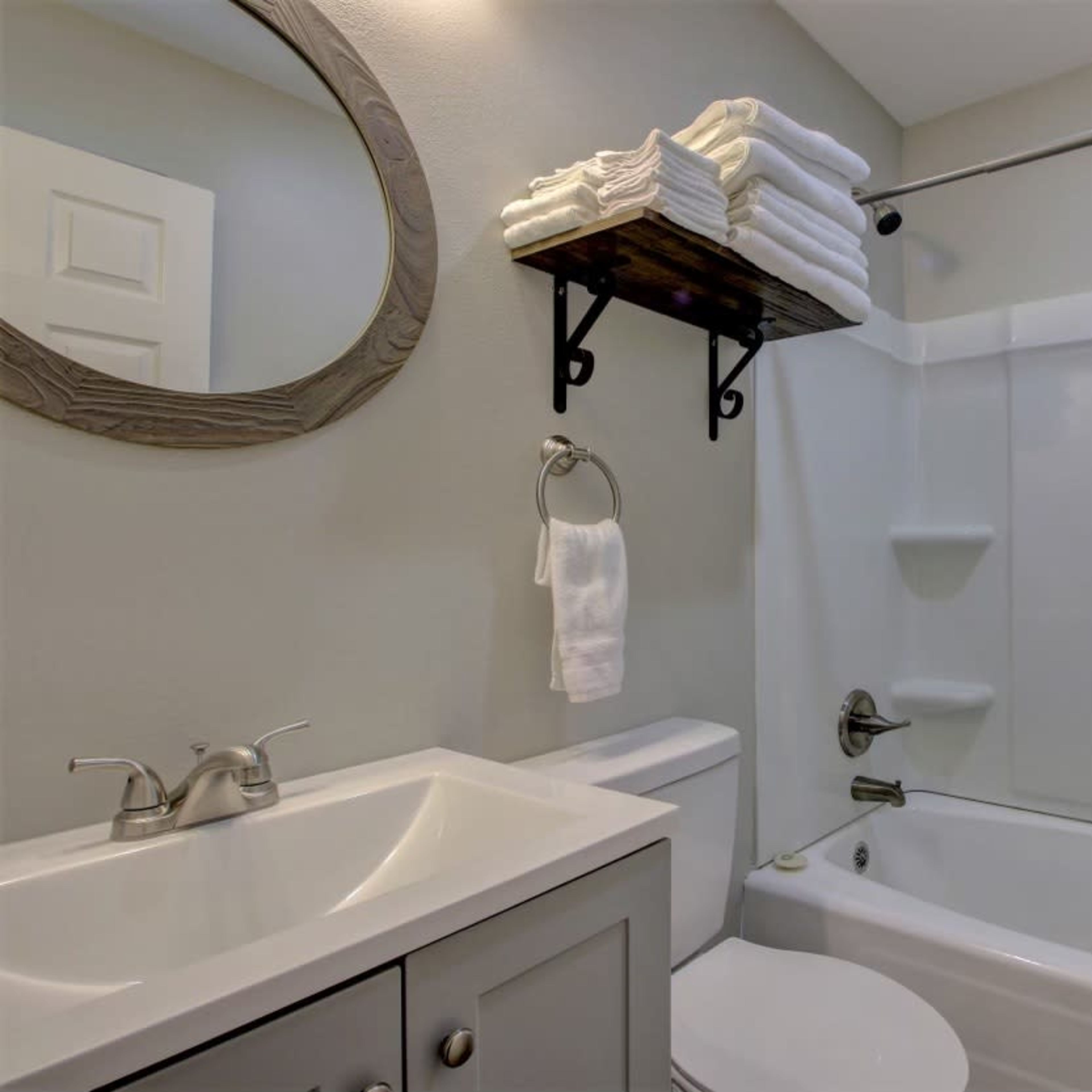 A bathroom with a shower curtain, a mirror, a sink with a cabinet, and neatly stacked towels on a shelf.