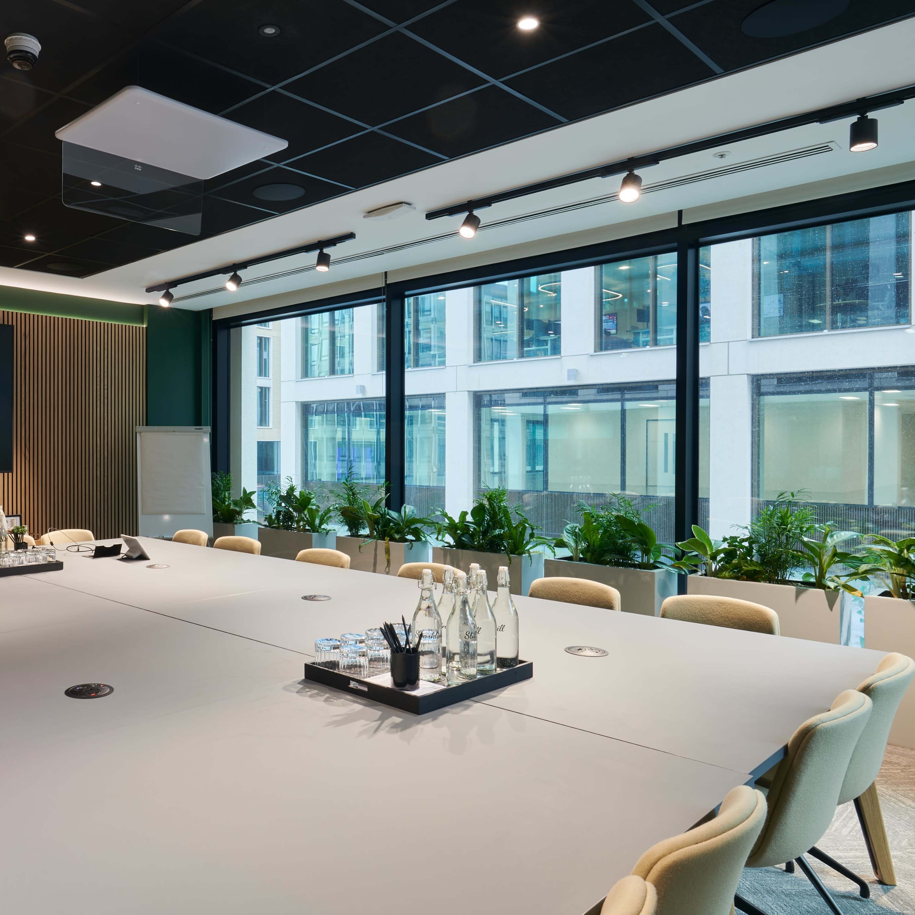 A modern conference room features a large table surrounded by chairs, with a screen on the wall and windows overlooking a cityscape.
