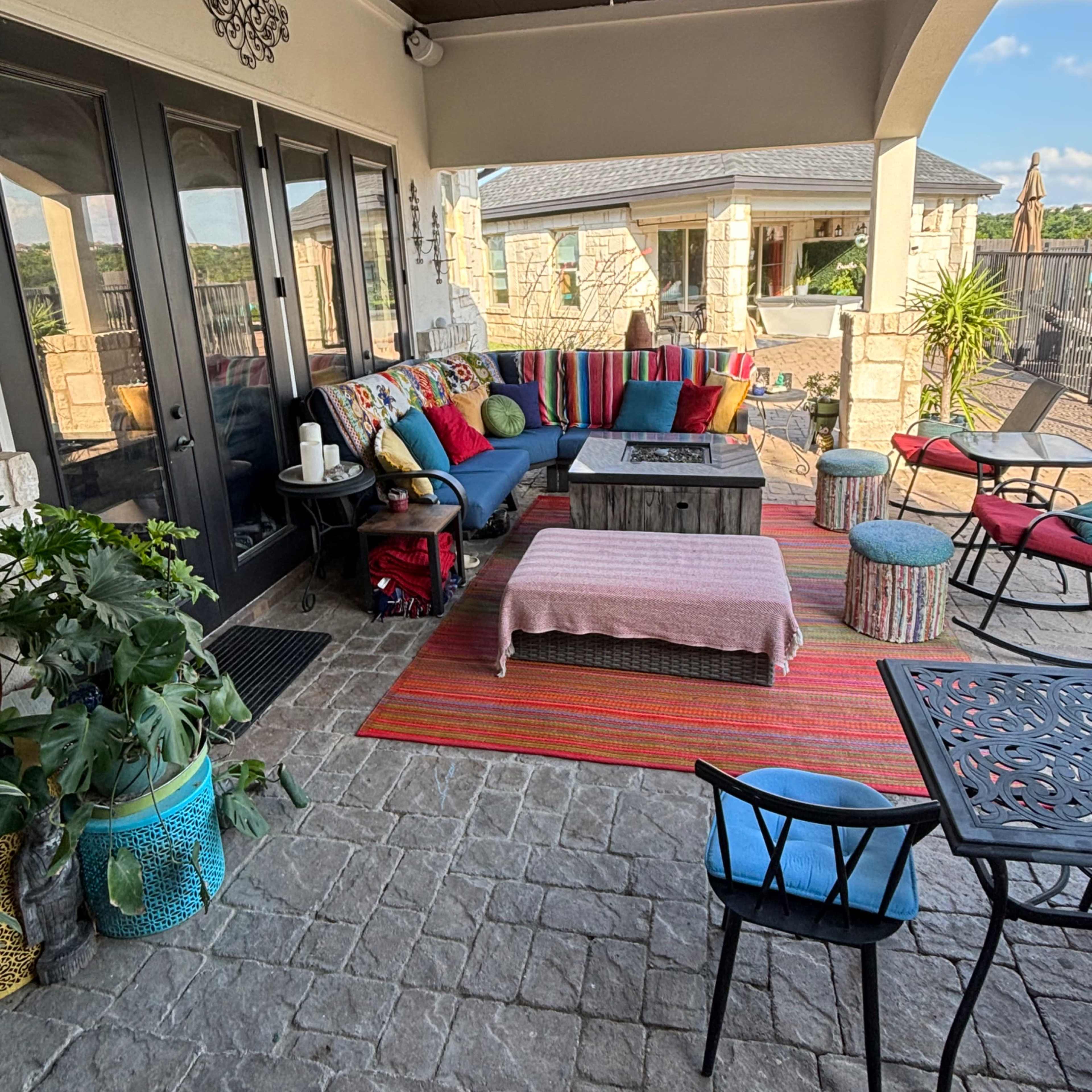 A stylish outdoor patio area furnished with colorful cushions, a wooden coffee table, and various seating options, surrounded by greenery and a view of the landscape.