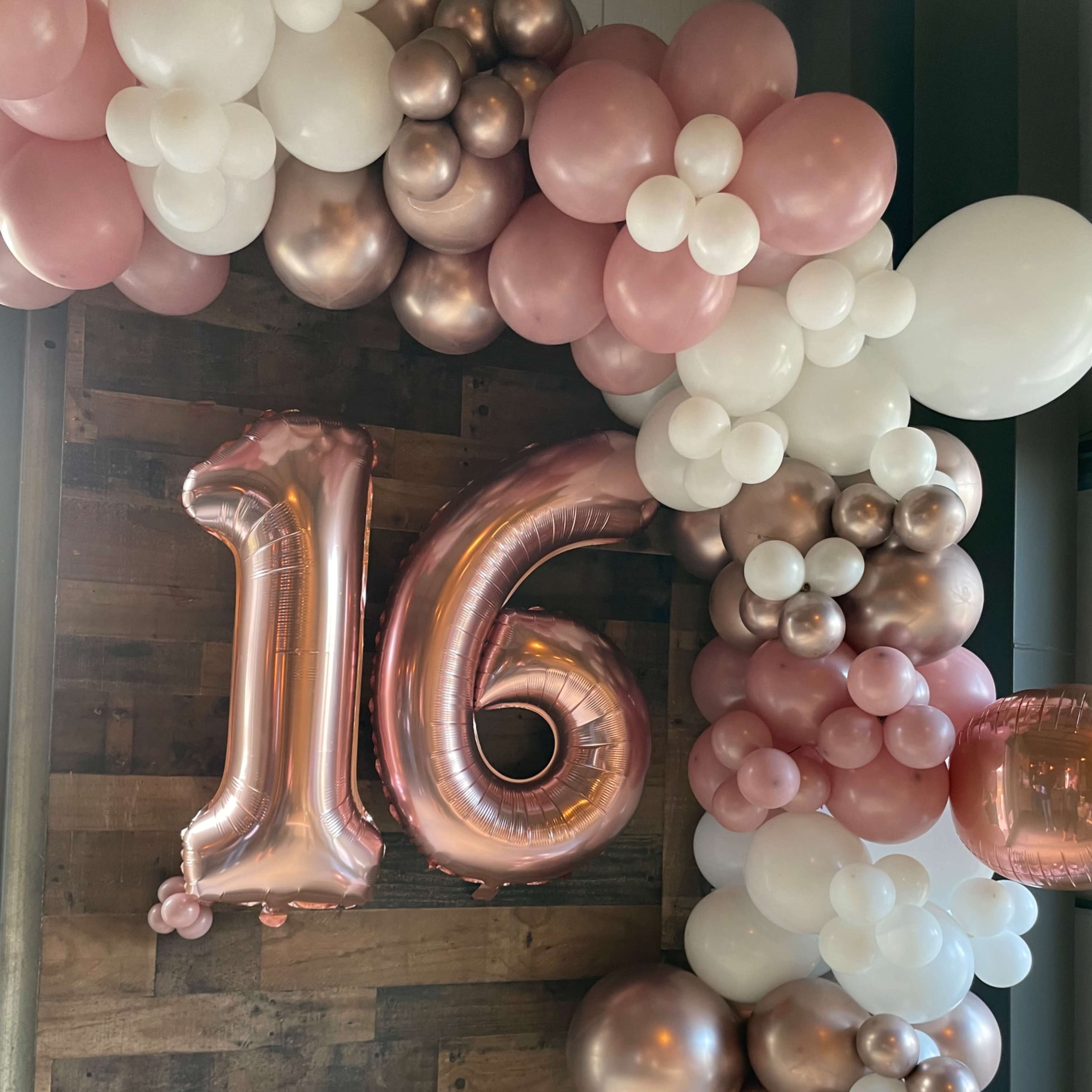 A decorative balloon arch with a large "16" balloon, incorporating shades of pink, white, and metallic colors against a wooden backdrop.