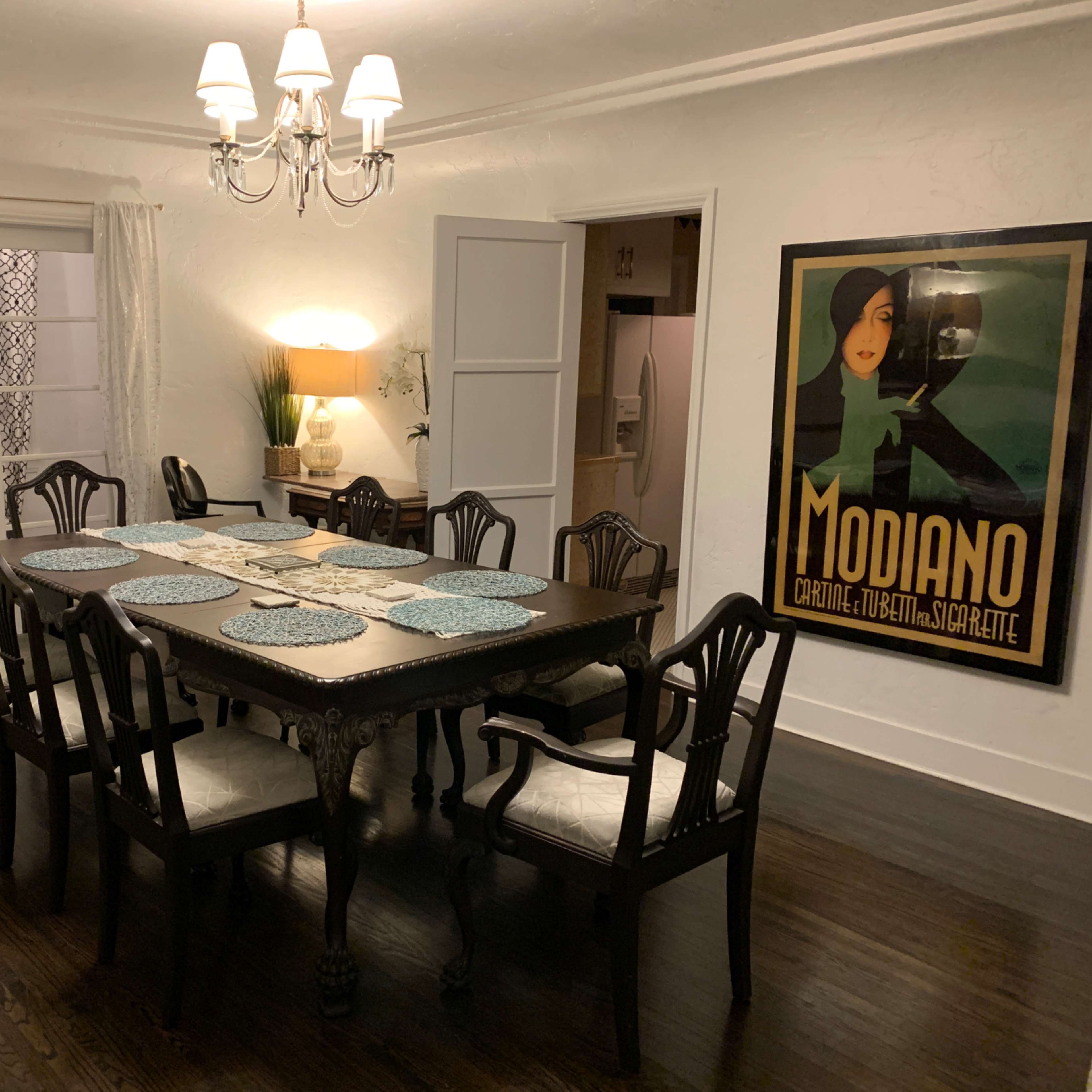 A large dining table with eight chairs is set for dinner in a well-lit room featuring a vintage poster on the wall and white curtains.