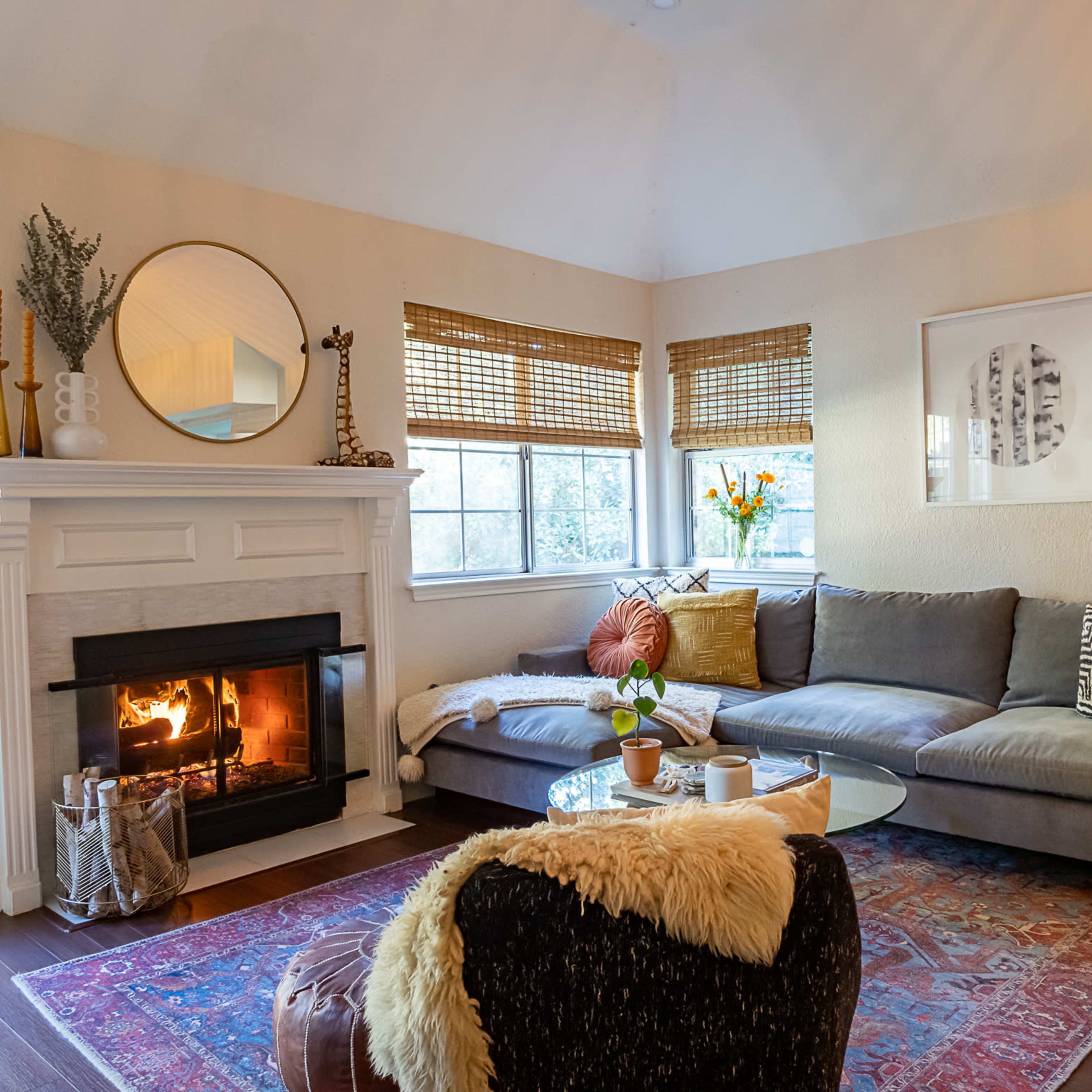 A cozy living room with a sectional sofa, a fireplace with a fire burning, and decorative elements like a round mirror and potted plants.