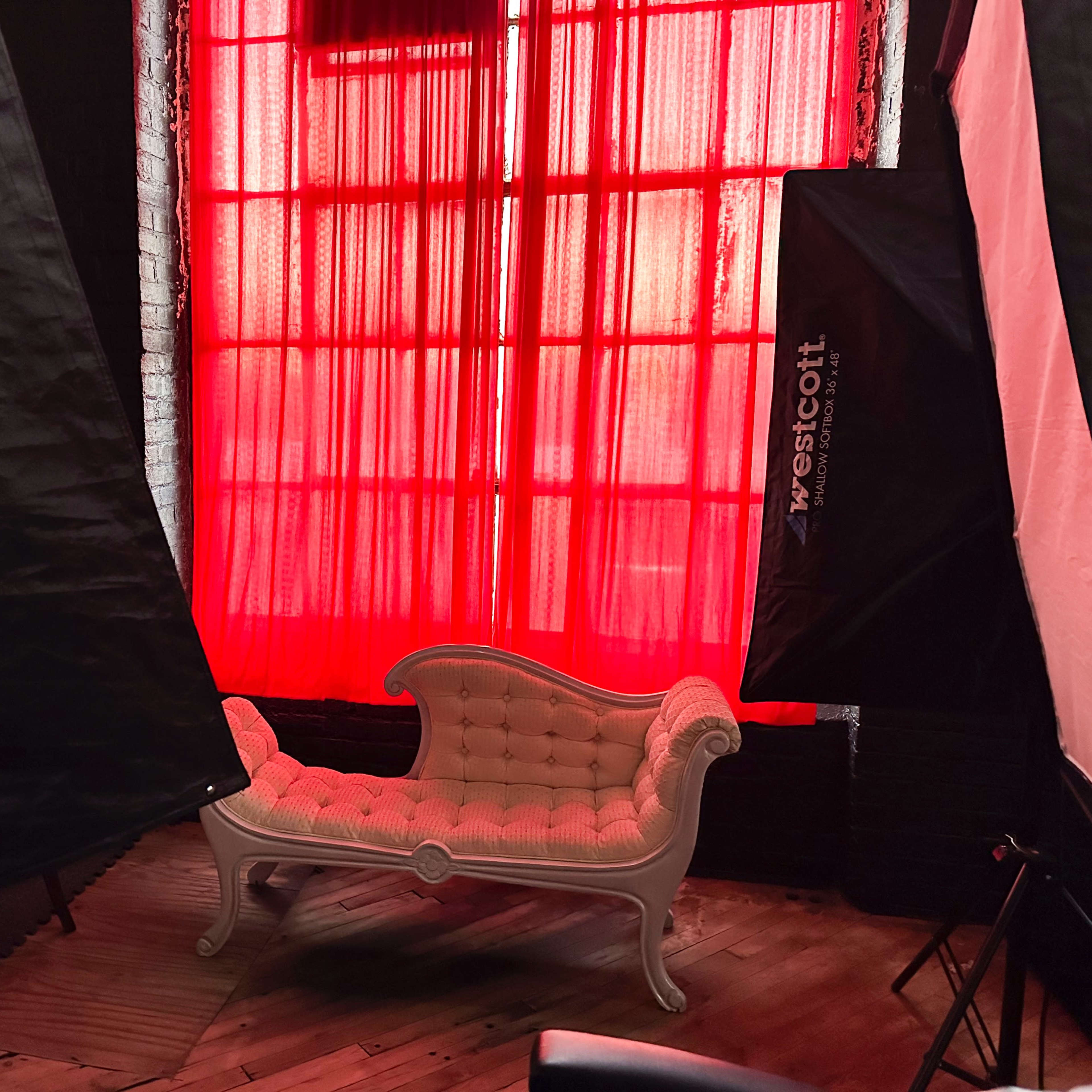 A vintage-style chaise lounge placed in front of large red curtains, surrounded by photography lighting equipment.