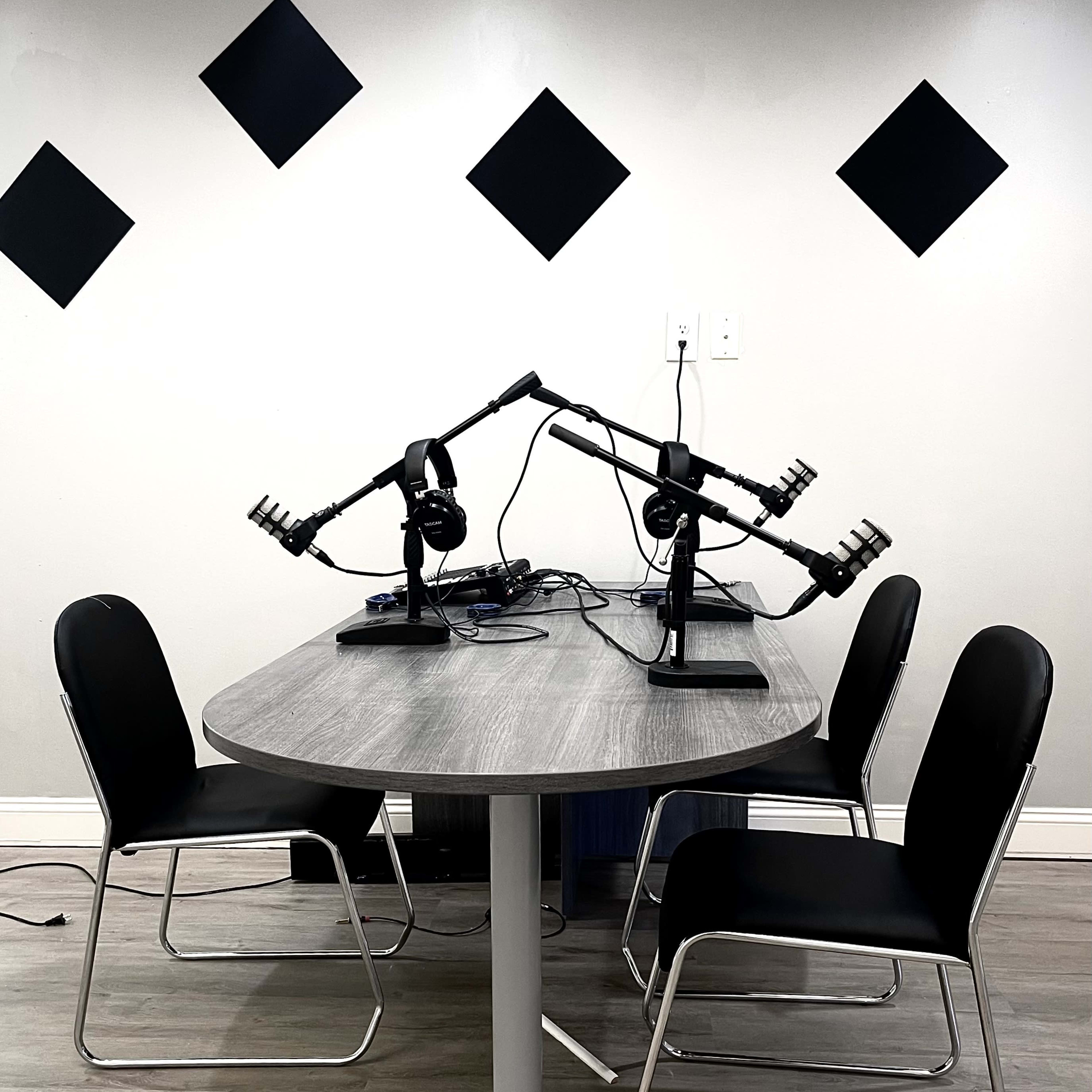 A round table with four black chairs, two microphone stands with microphones, and headphones arranged on top, set against a plain wall with black diamond shapes.