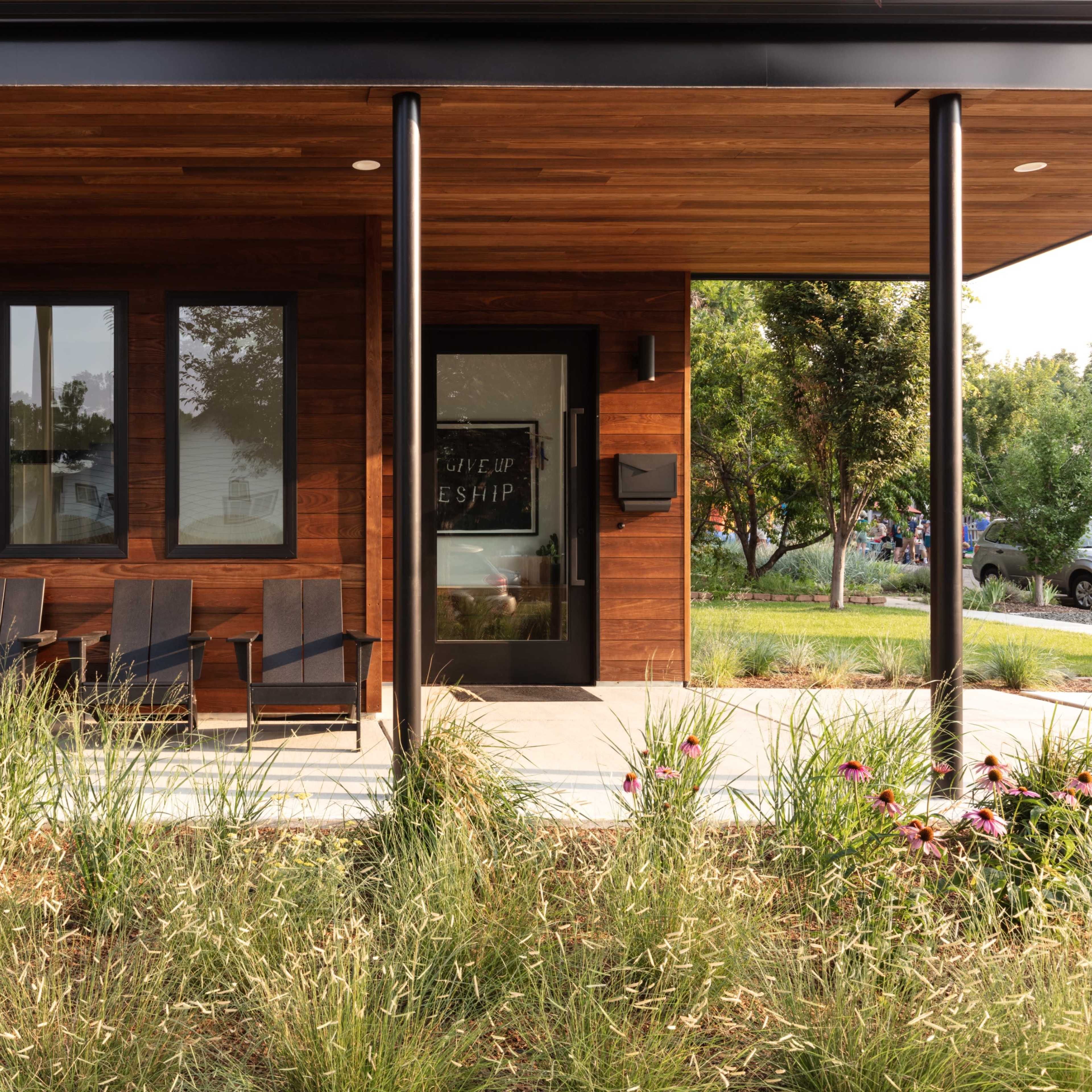 A modern house with a wooden facade, featuring a porch with three black Adirondack chairs and a landscaped yard.