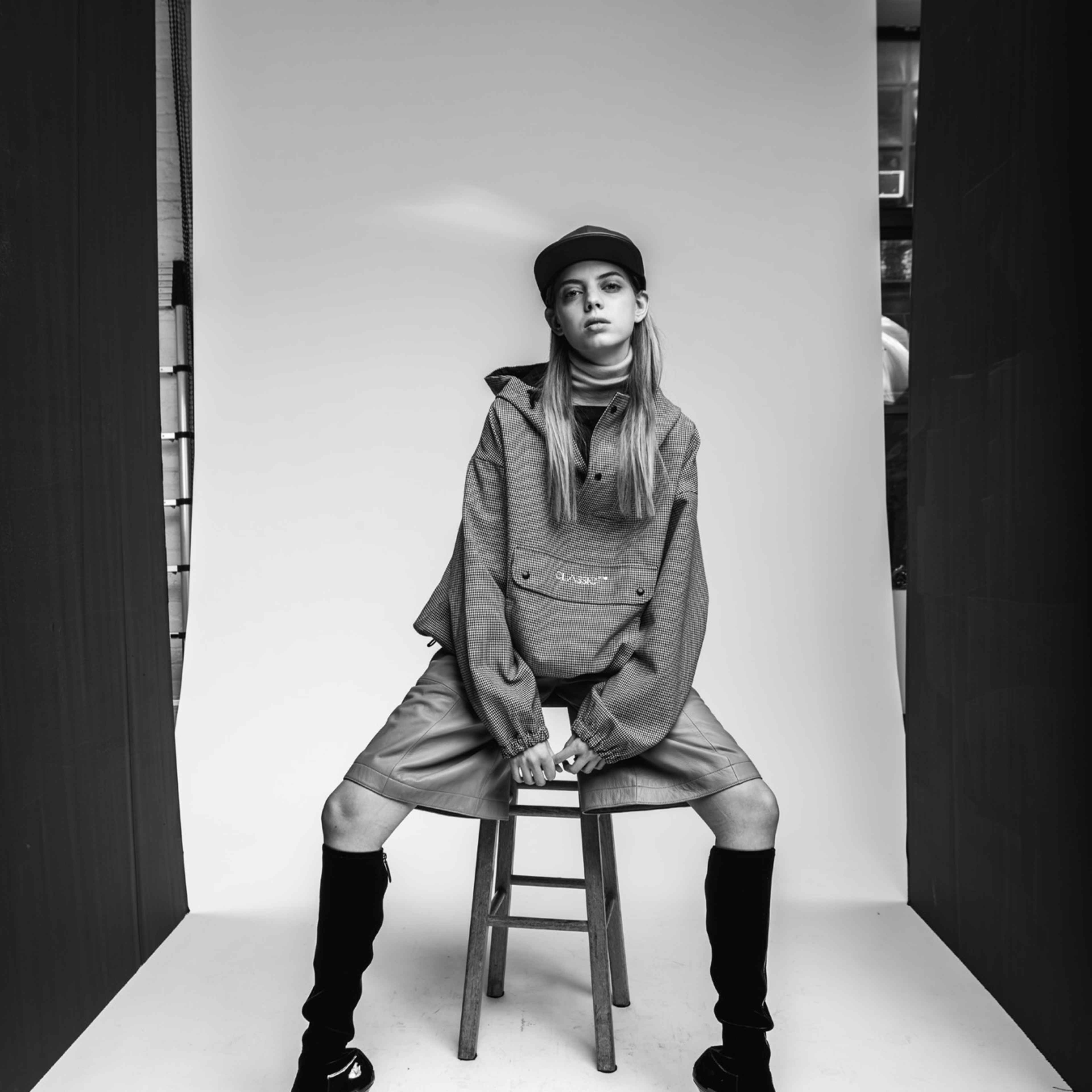 A model in stylish attire sits on a stool against a plain backdrop in a black-and-white photo.