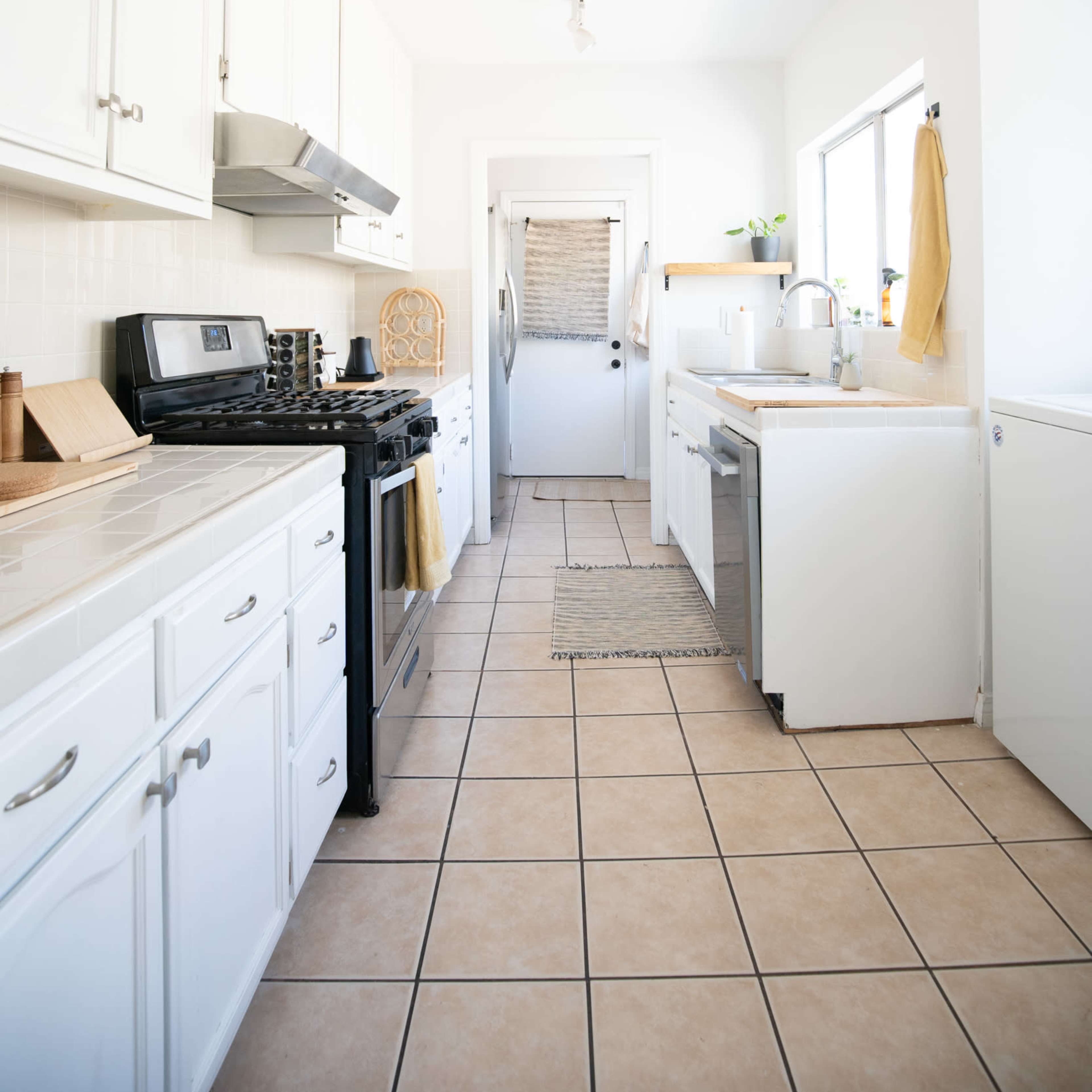 A long, narrow kitchen with white cabinetry, a gas stove, stainless steel appliances, and beige tiled flooring.
