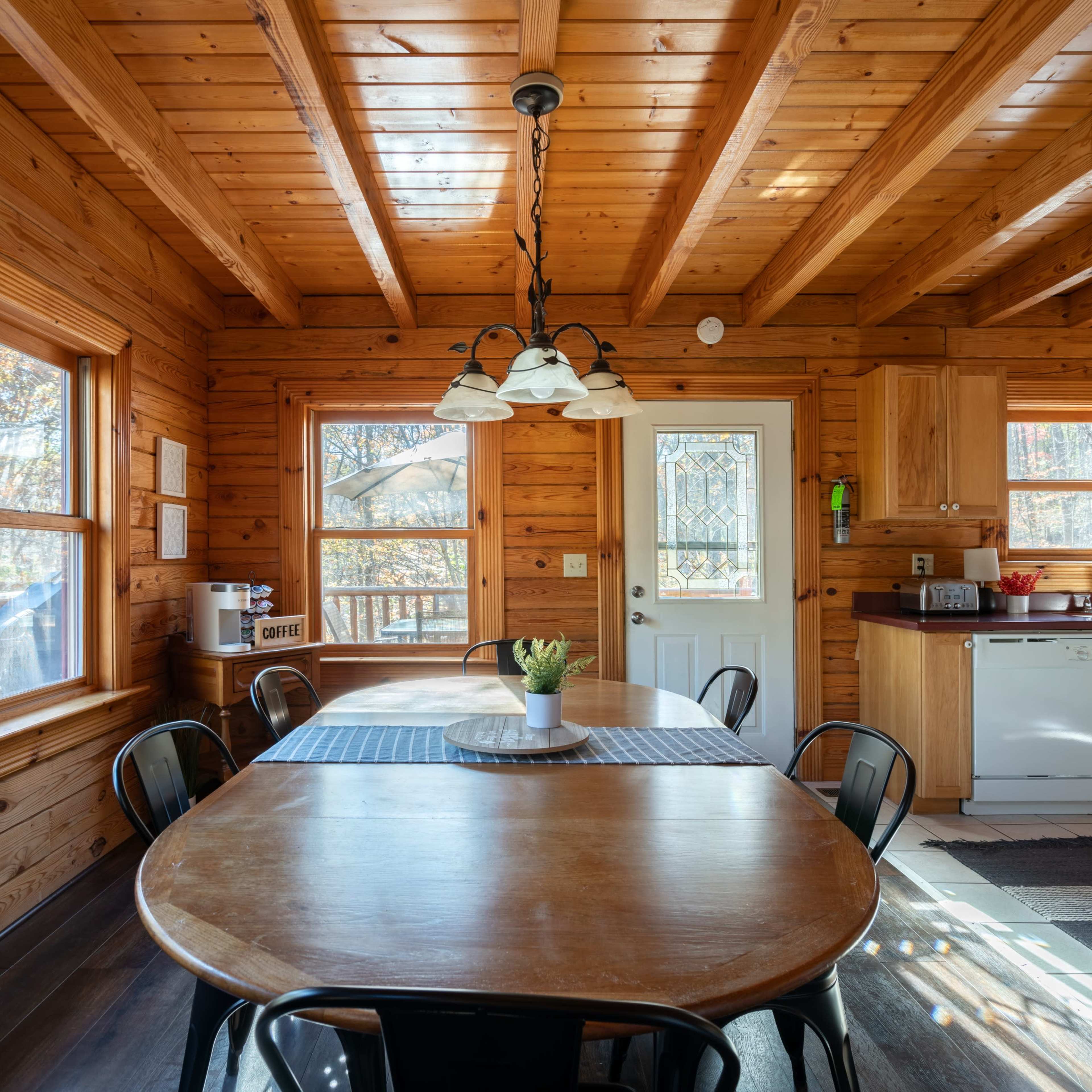 A wooden dining area features a large table surrounded by black chairs, with windows showcasing a view of the outdoors.