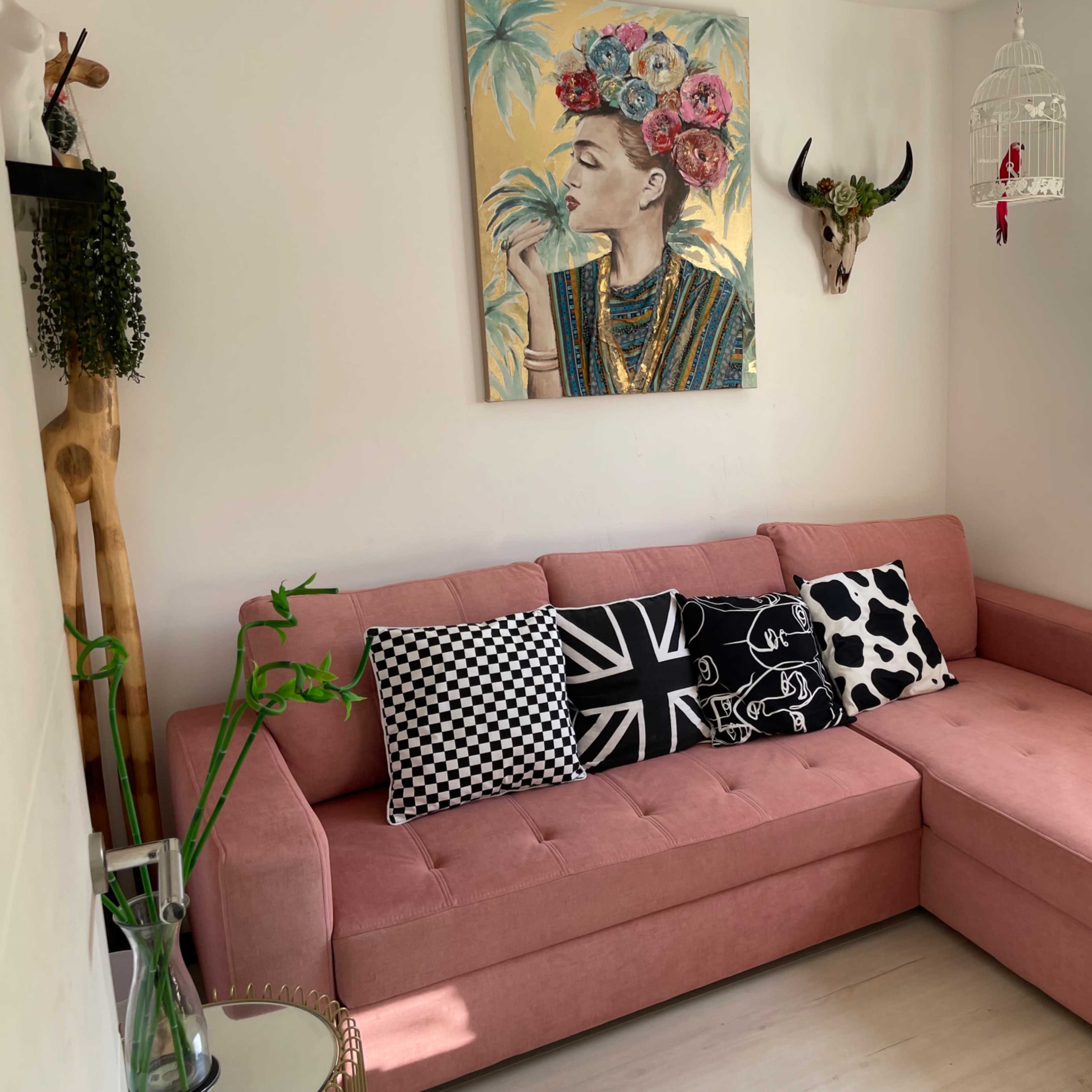 A corner of a room featuring a pink sectional sofa adorned with patterned cushions, a large artwork on the wall, and decorative items including a mounted skull and a hanging birdcage.