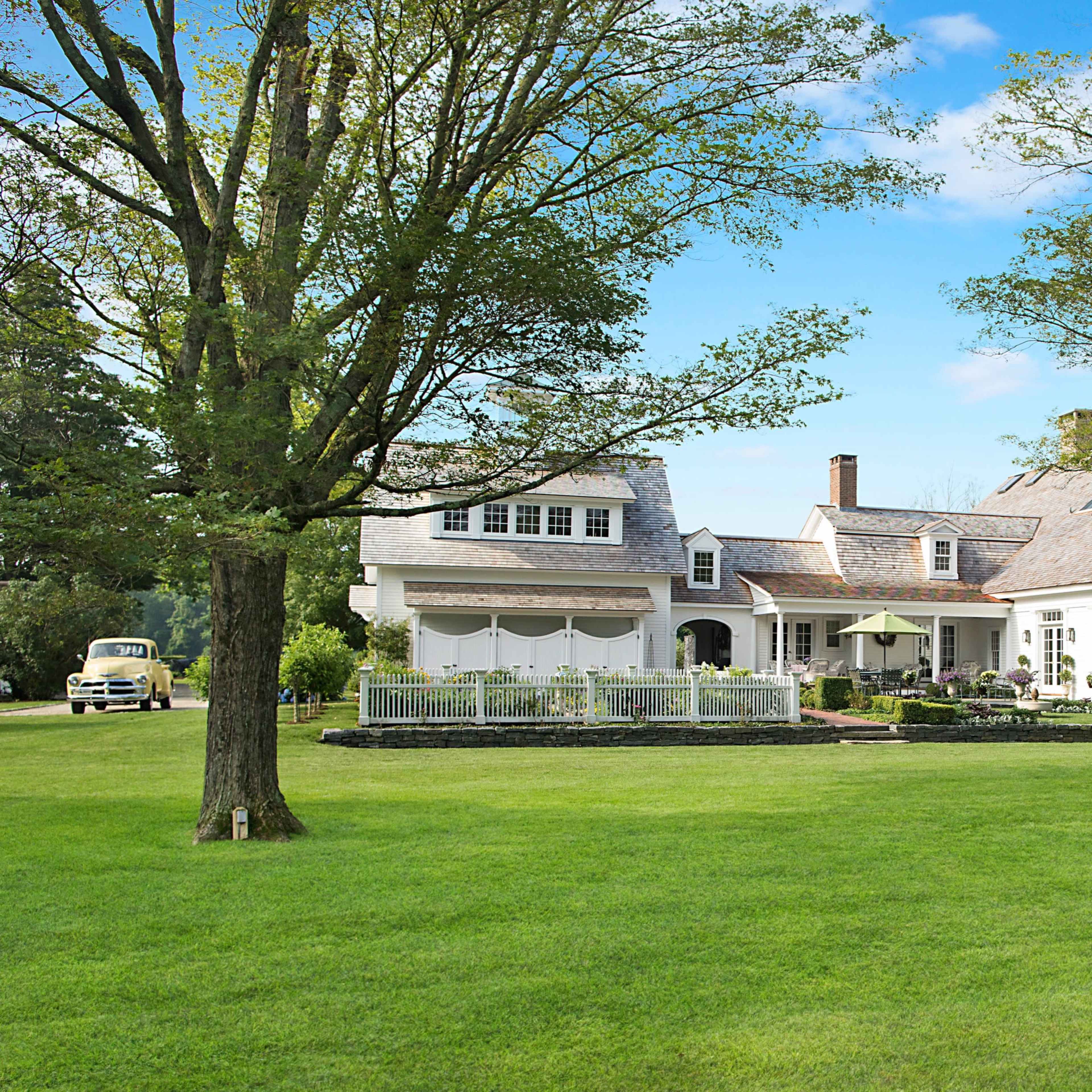 A large, white two-story house is surrounded by a lush green lawn and tall trees, with a stone walkway leading to the entrance.