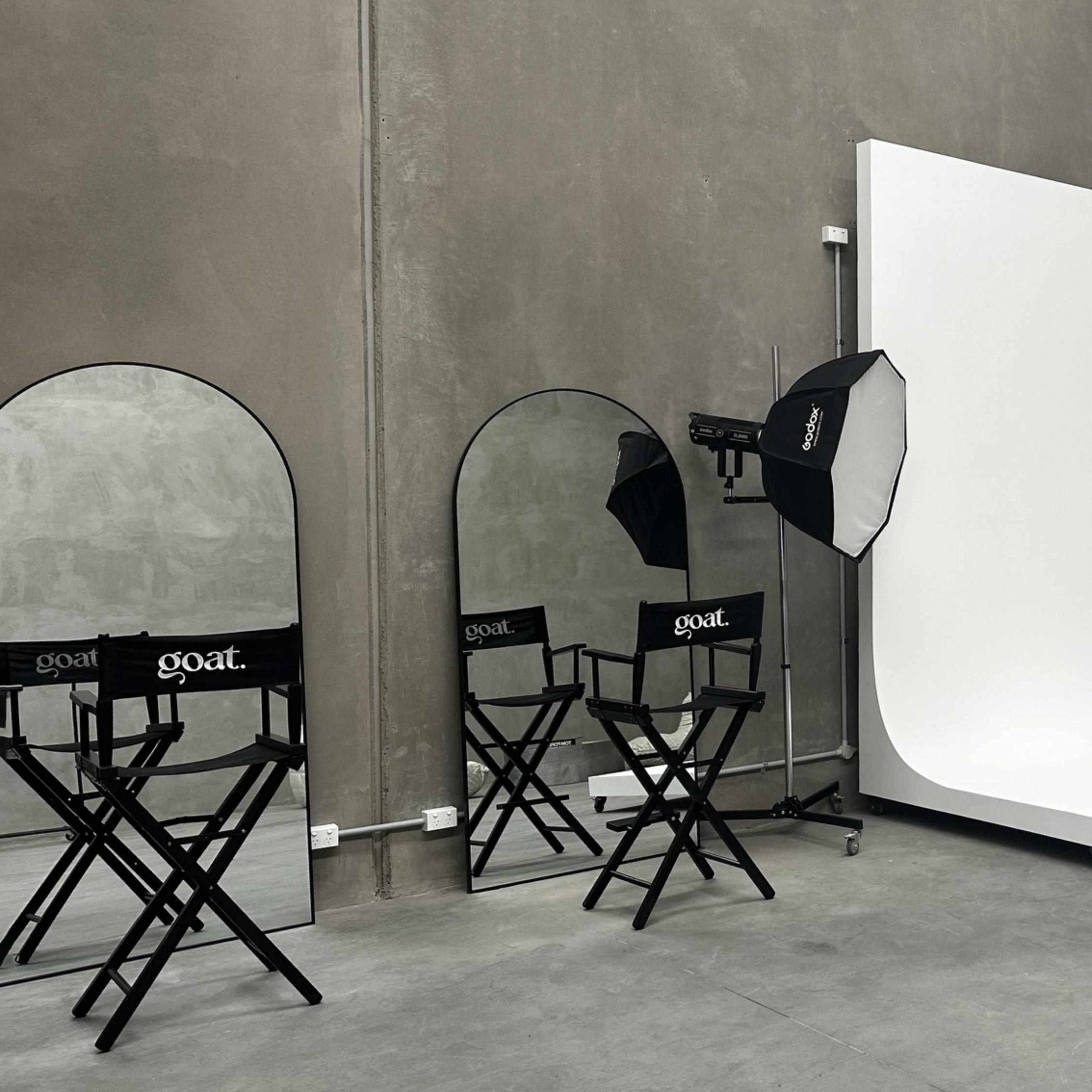 A minimalist photography studio with a white backdrop, two softbox lights, and two director's chairs labeled "goat."