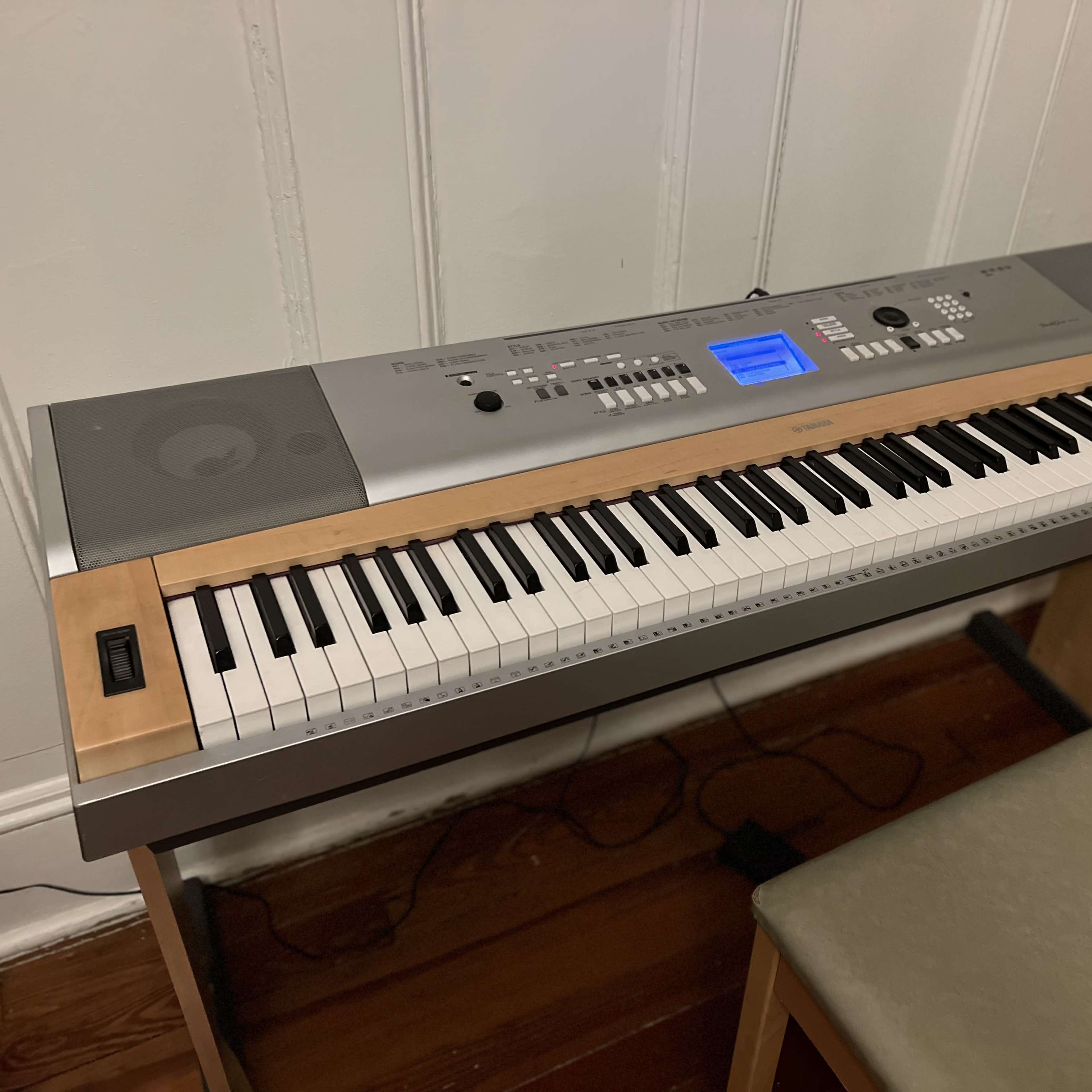 A digital piano with a wooden finish on its front and a control panel displaying blue lights, set on a wooden floor next to a chair.