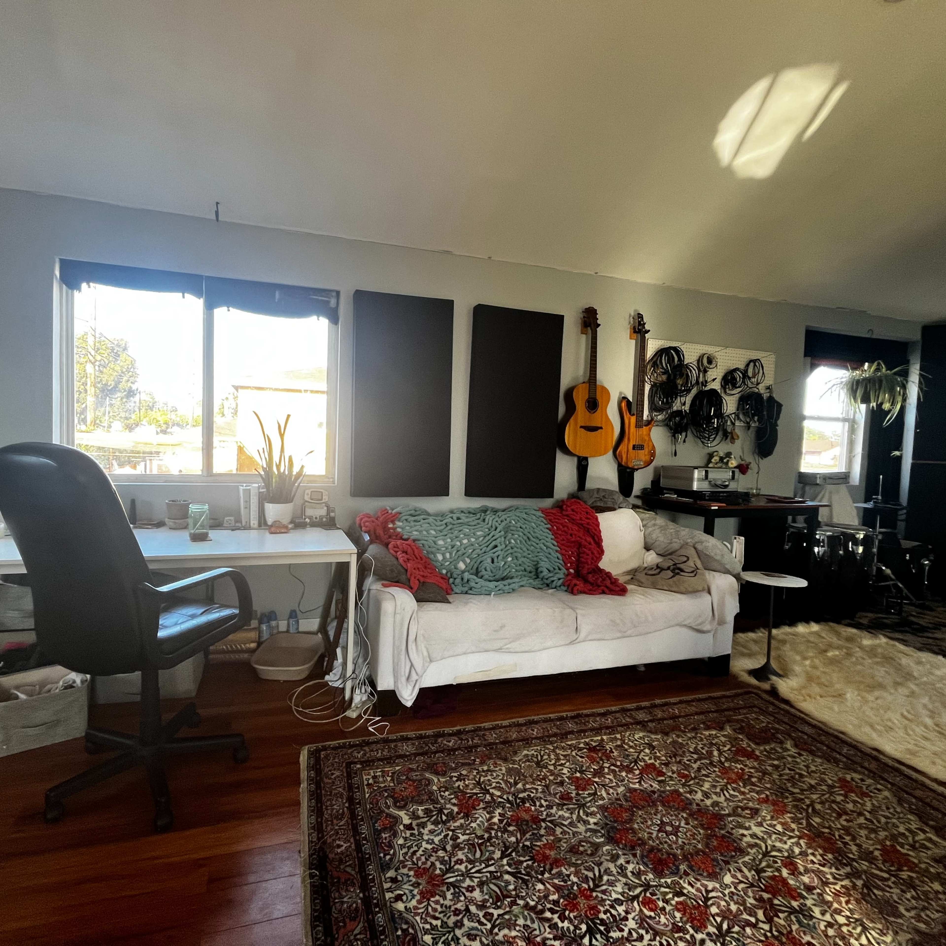 A spacious room with a white couch, a workspace with a desk and a chair, and musical instruments displayed on the wall.