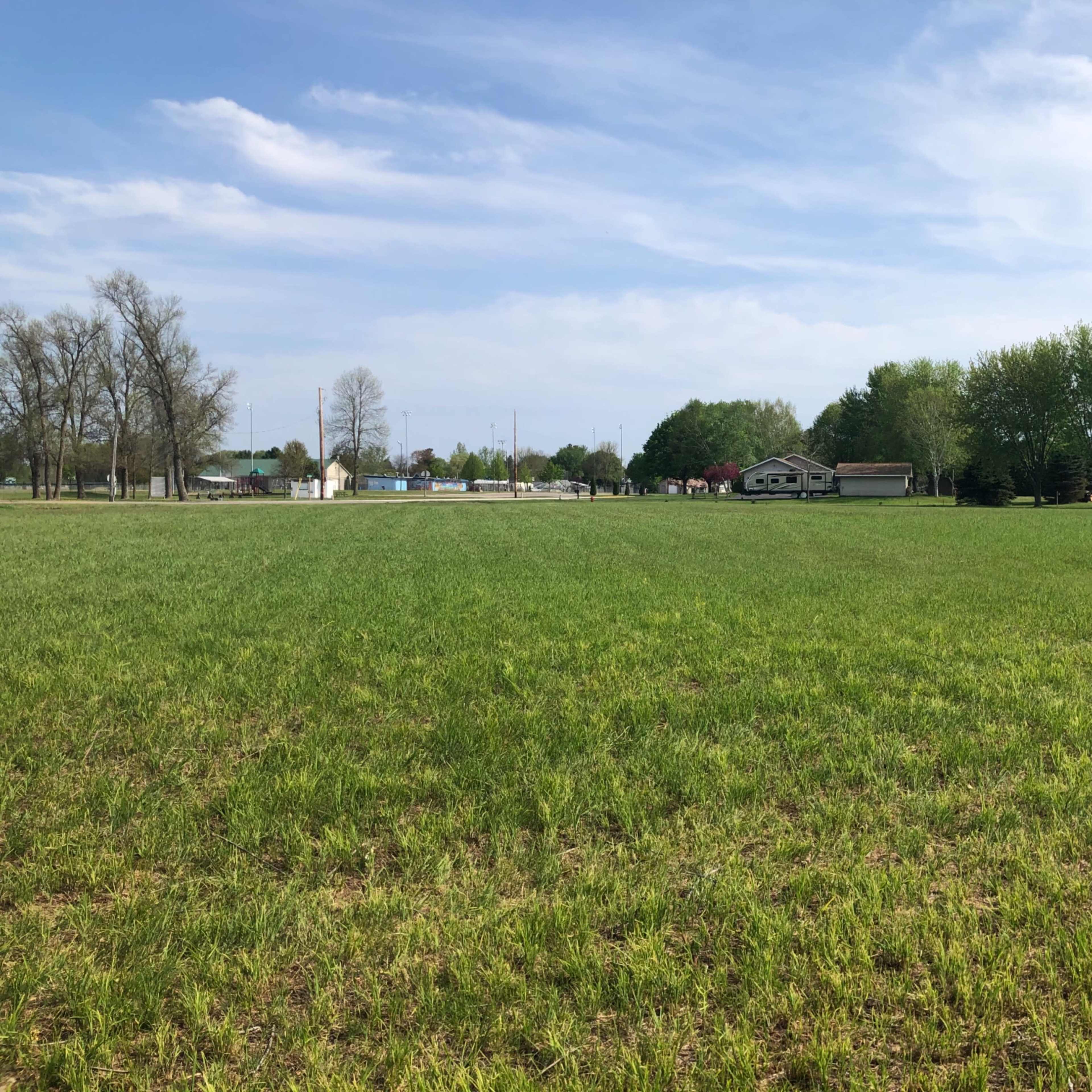 A wide, grassy field extends towards a line of trees and a small cluster of buildings in the background under a blue sky.