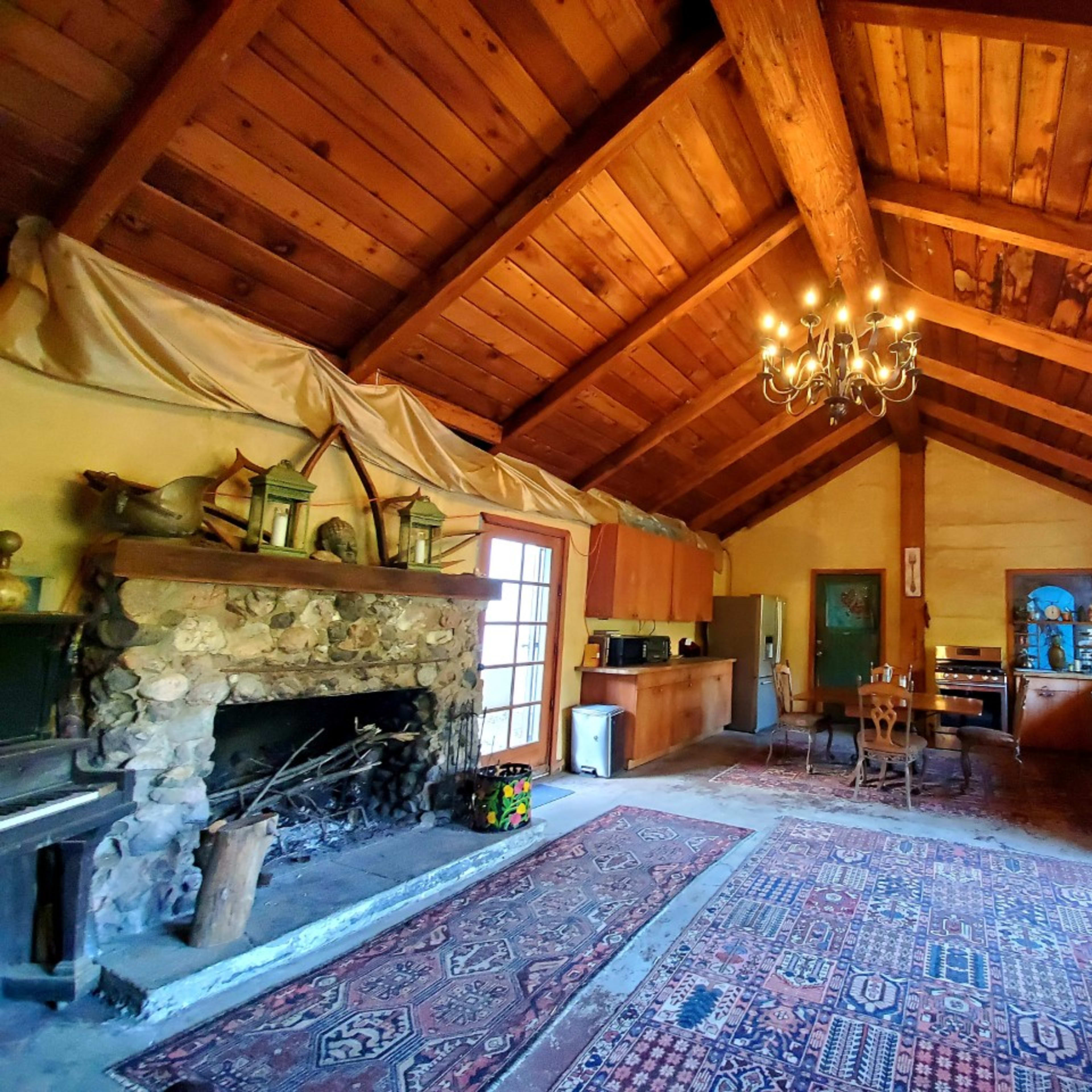 The interior of a rustic cabin features exposed wooden beams, a stone fireplace, and a large area rug on a wooden floor.