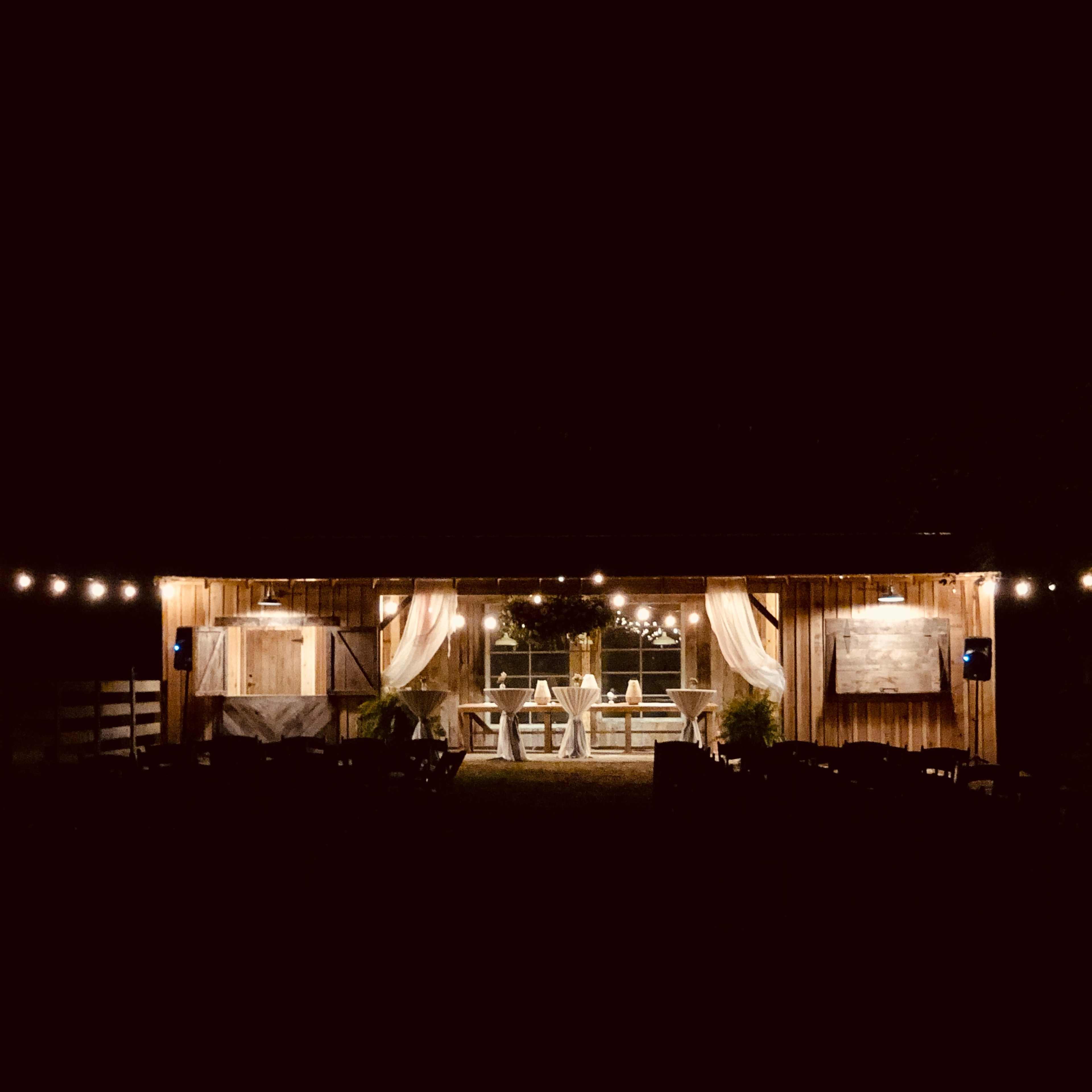 An illuminated outdoor gathering area with string lights, set up with tables and chairs in a rustic wooden structure at night.