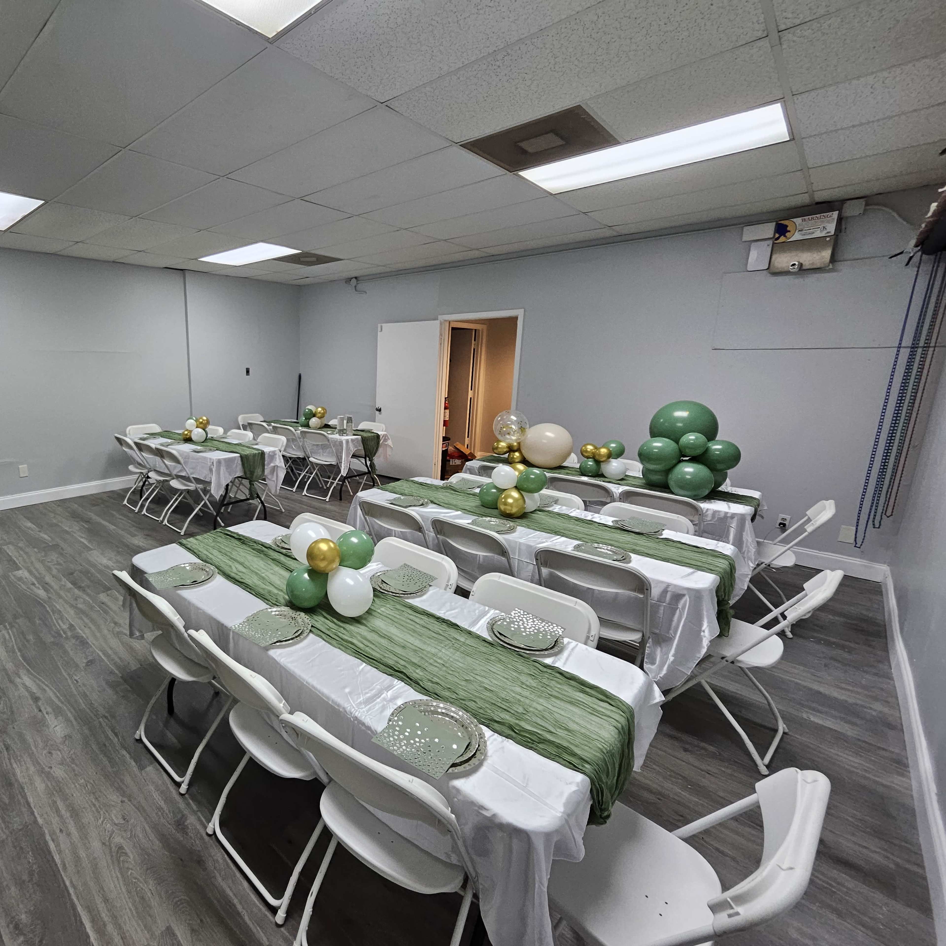 A spacious event room is arranged with long tables covered in white cloth and green table runners, surrounded by white folding chairs and decorated with balloon clusters in various colors.