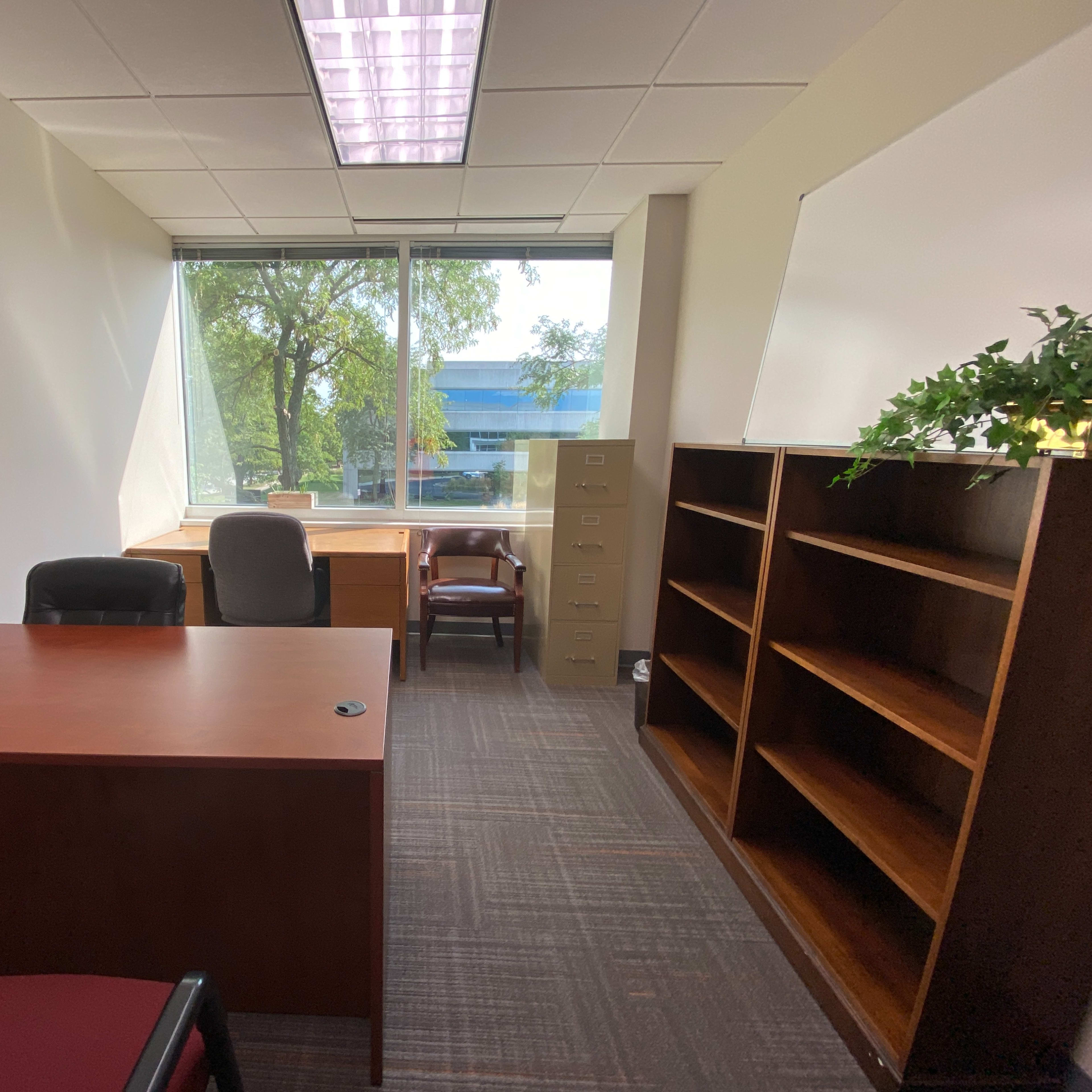 An office with a wooden desk, an office chair, a filing cabinet, and shelves, all positioned near a window that offers natural light.