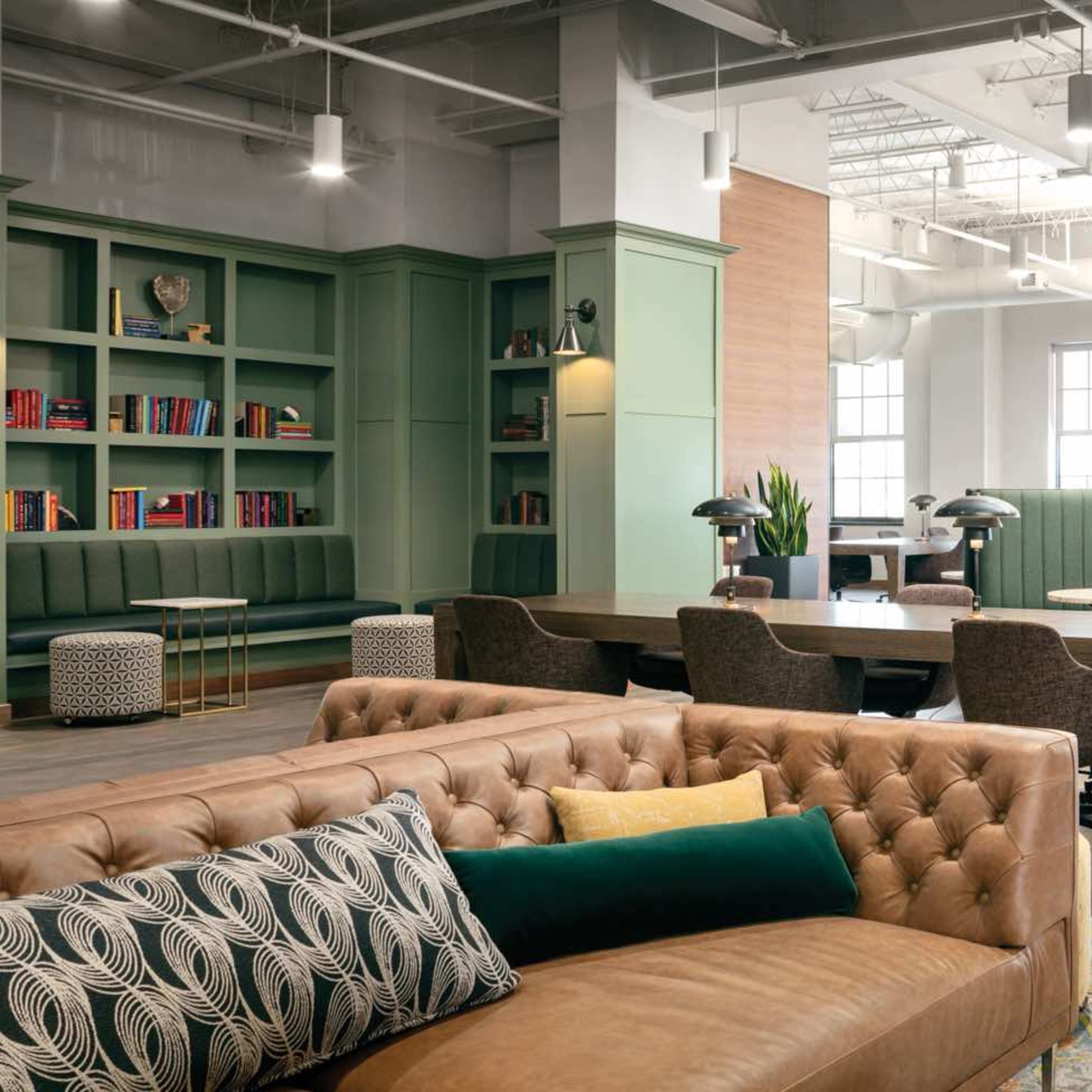 A modern office space with green shelving filled with books, a brown leather couch, a large wooden table, and seating areas separated by partitions.