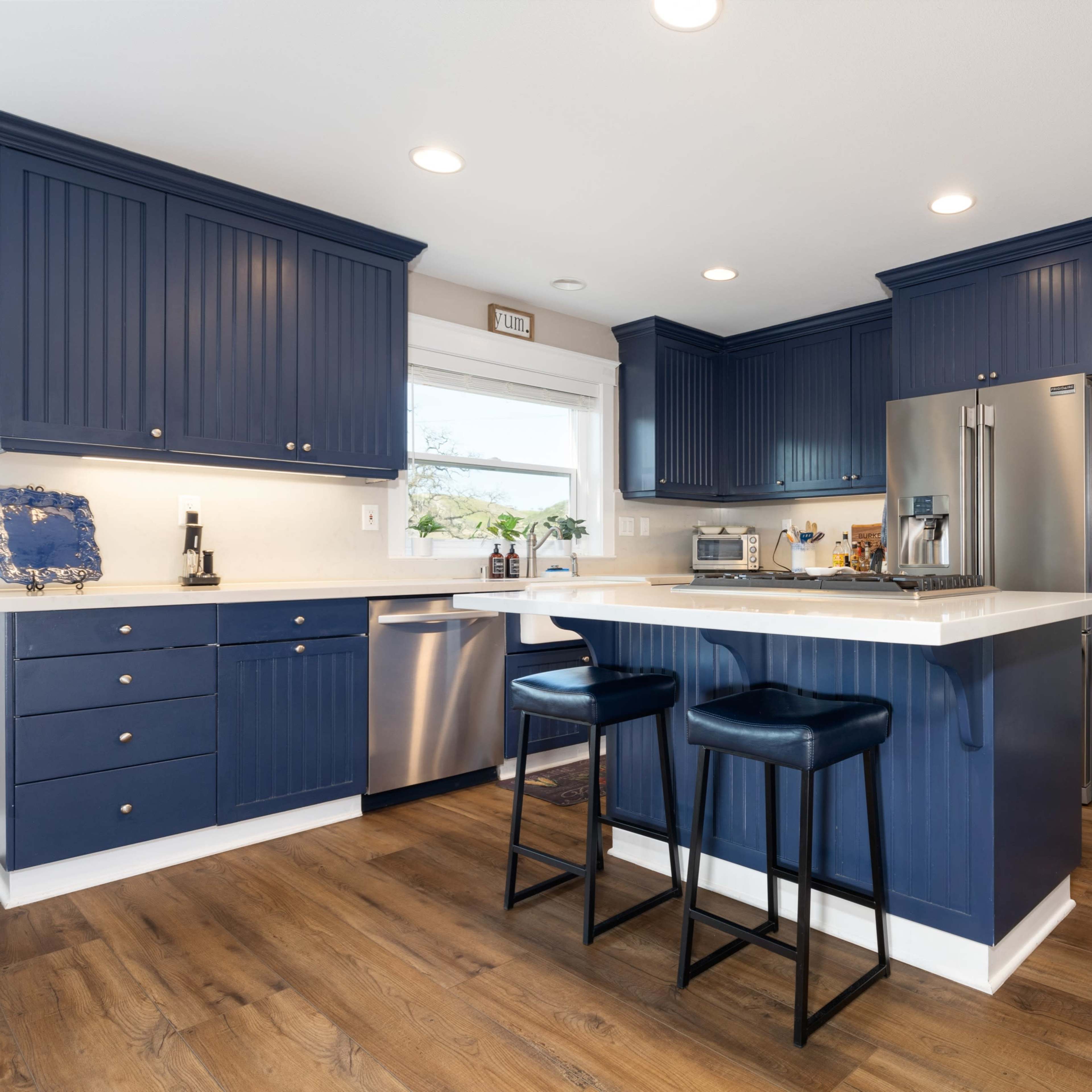 A modern kitchen with blue cabinetry, stainless steel appliances, and a light countertop, featuring two bar stools at a central island.