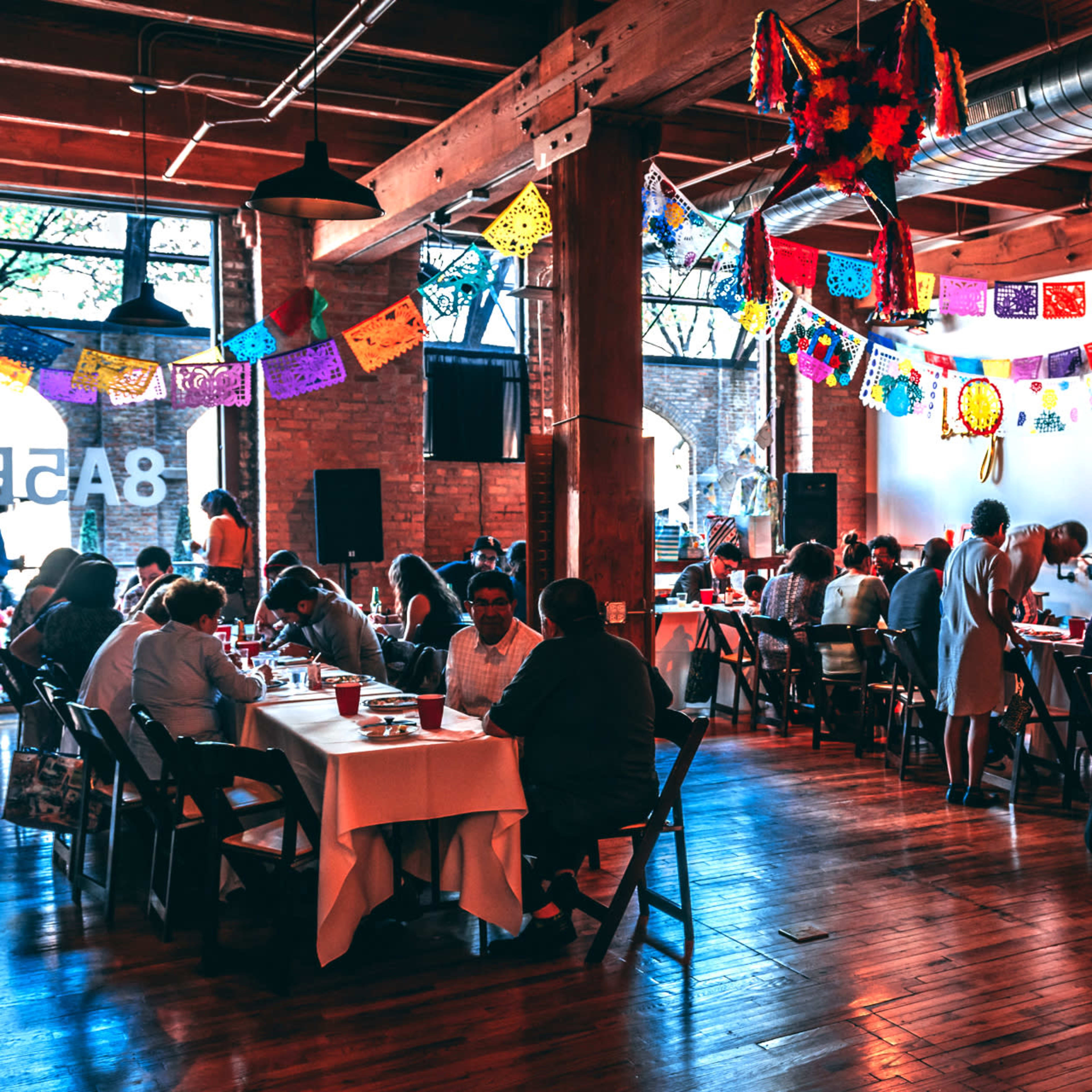 A busy indoor event space features attendees seated at tables, enjoying a meal under colorful decorations and lights.