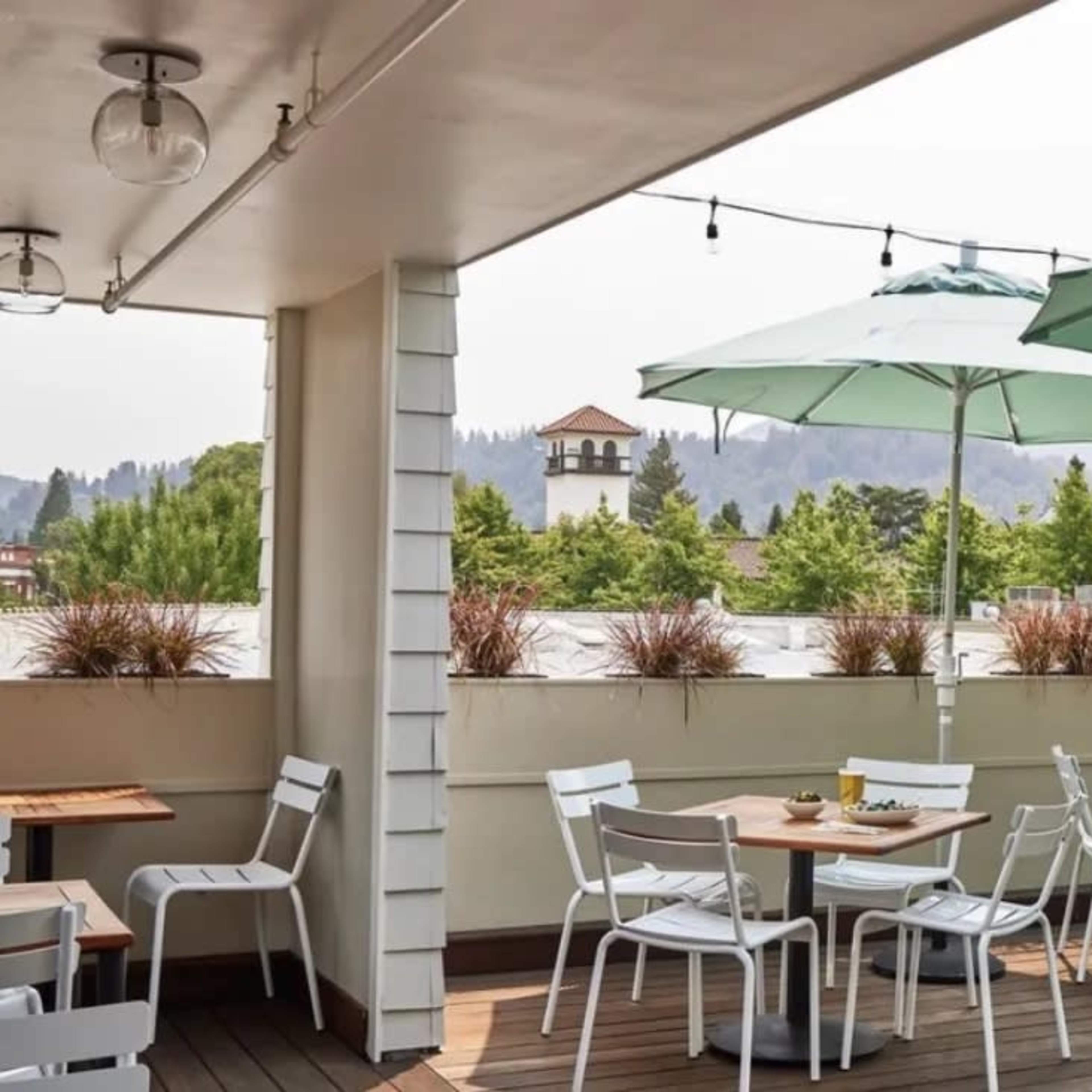 A rooftop patio features white chairs and tables under green umbrellas, surrounded by greenery and distant hills.