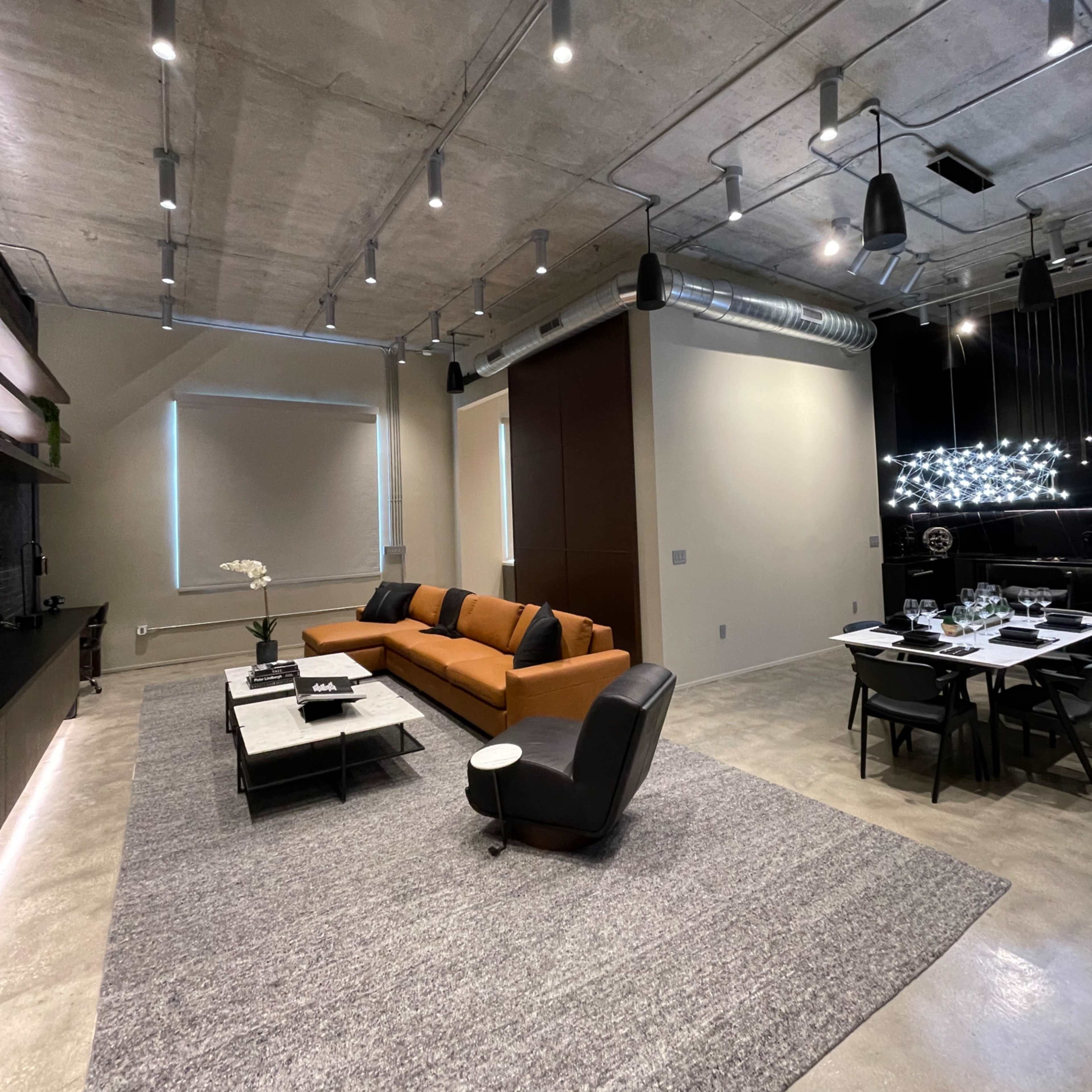 A modern living room with an orange sofa, a black chair, a coffee table, and a dining area illuminated by pendant lights.