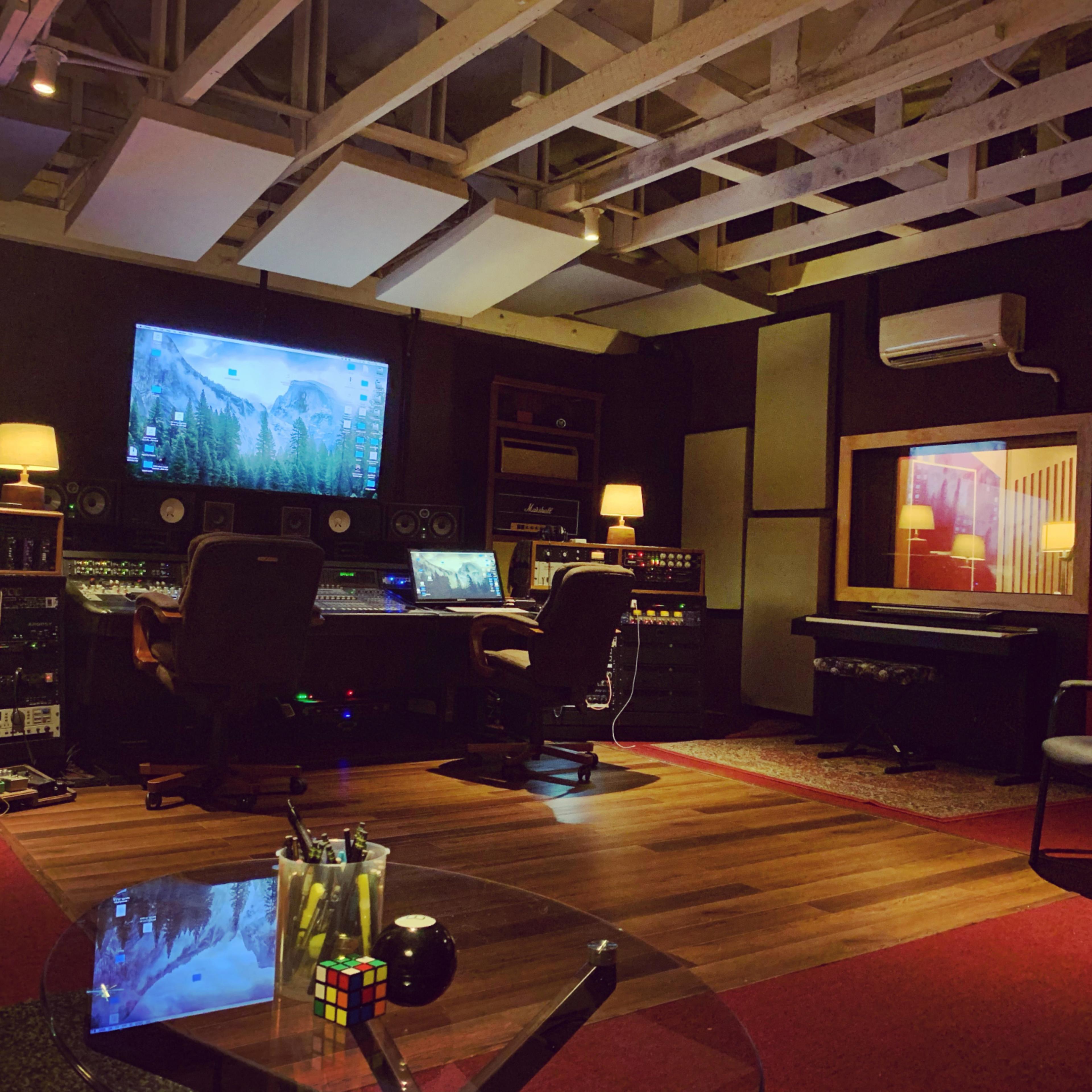 A music studio is equipped with a central mixing desk, monitors, and a variety of audio equipment, surrounded by seating and decorative lighting.
