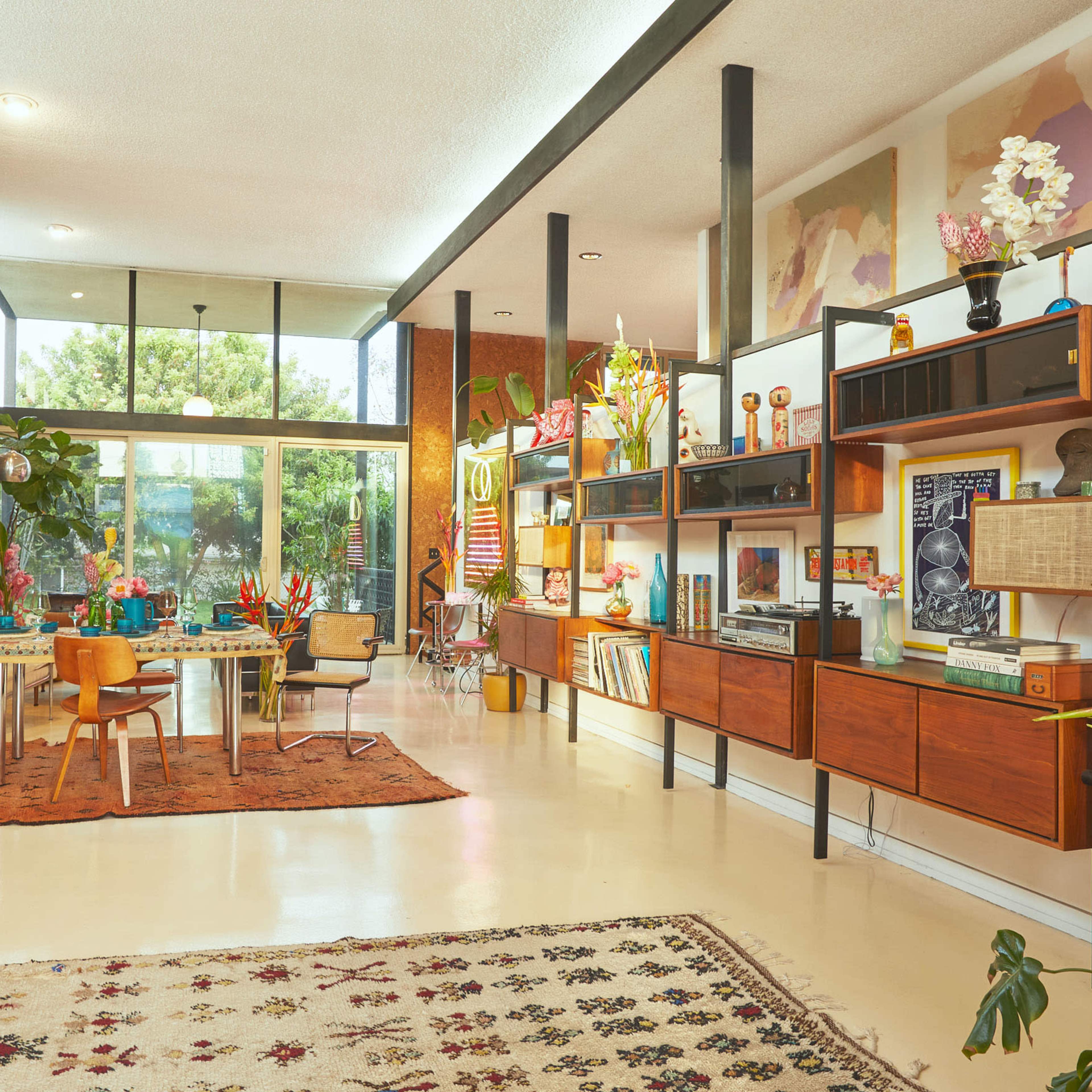 A modern living room features large windows, stylish furniture, and colorful decor, with plants and a dining area complementing the space.