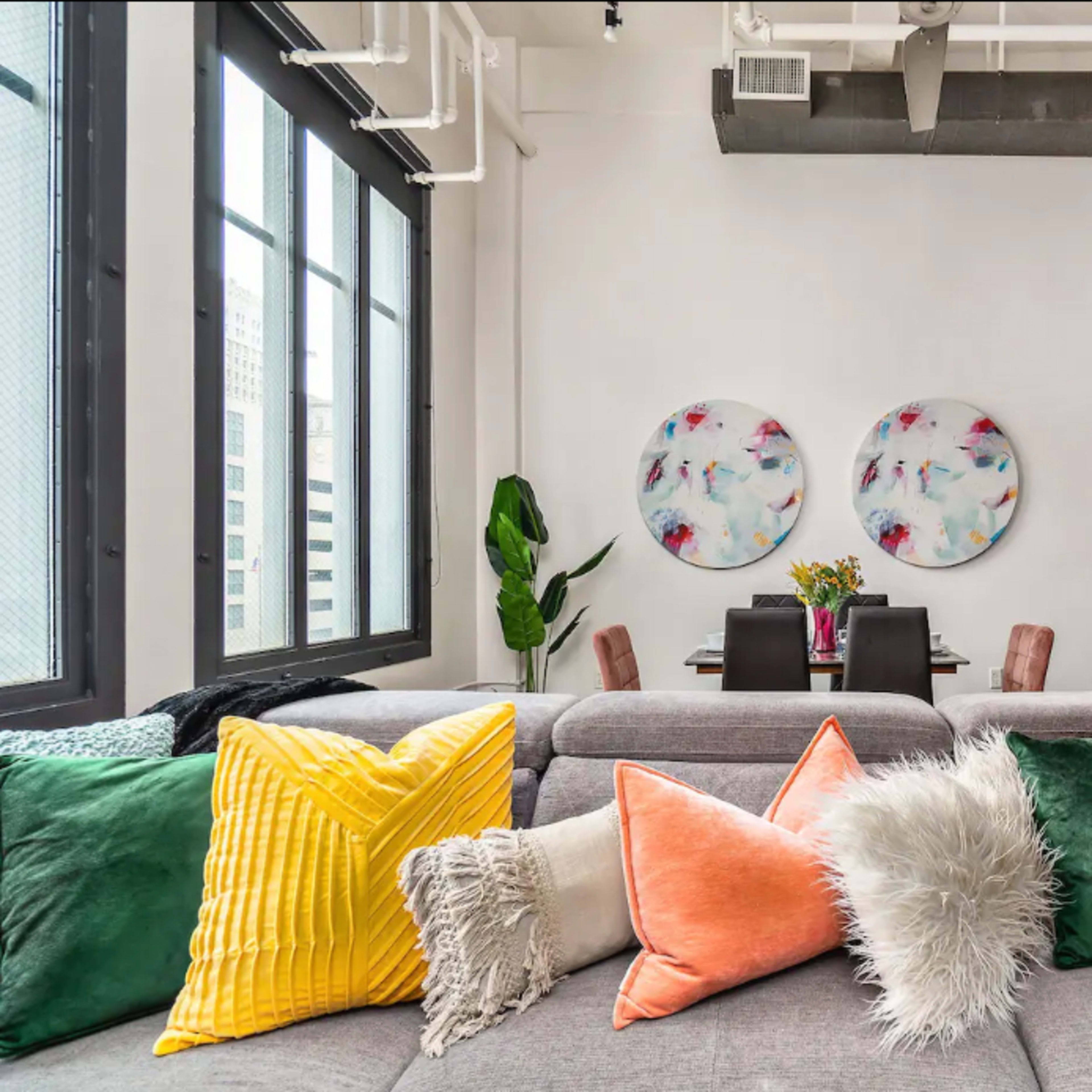 A modern living space featuring a gray sofa adorned with colorful decorative pillows, a dining area in the background, and large windows letting in natural light.