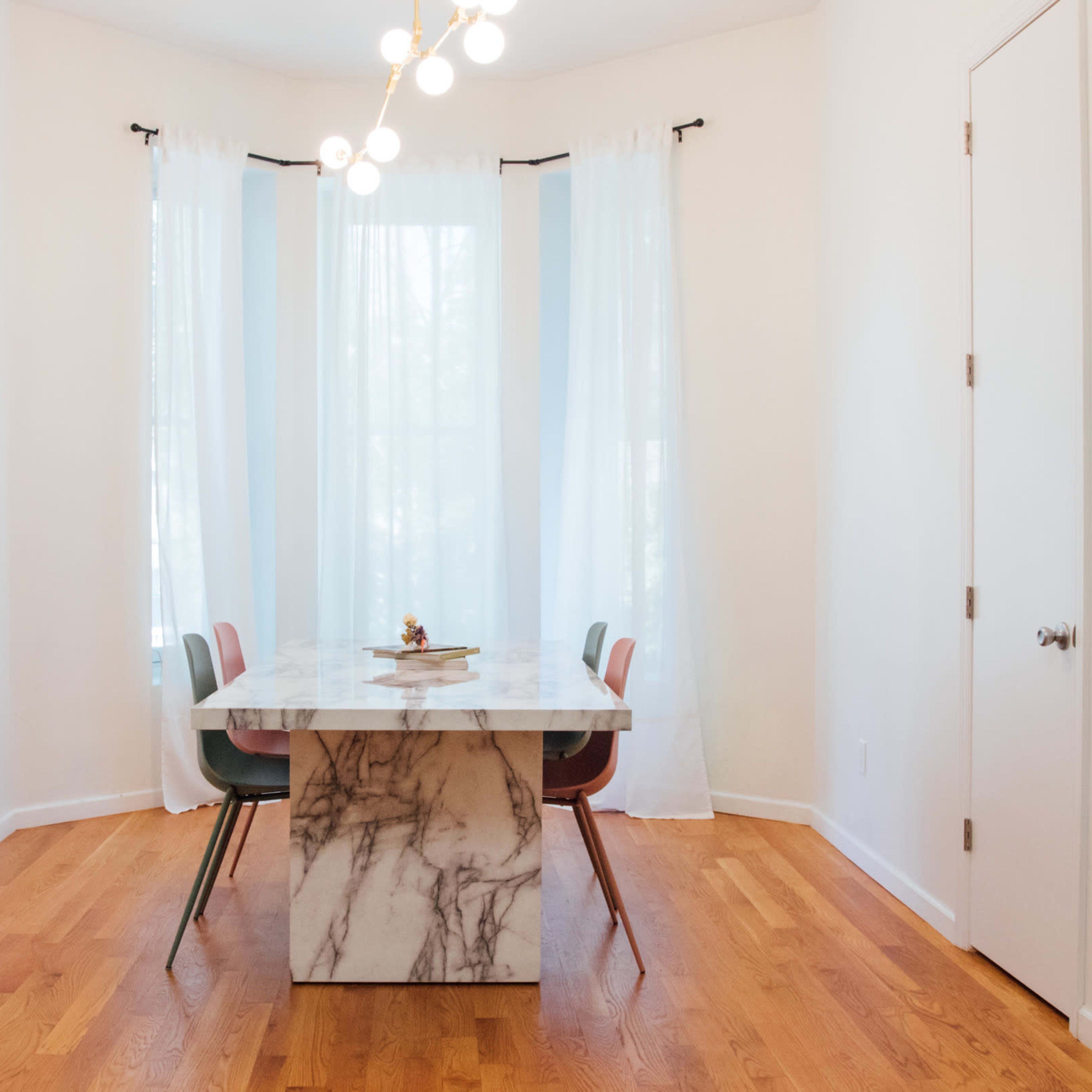 A modern dining area features a marble table surrounded by four colorful chairs, with large windows draped in sheer curtains.