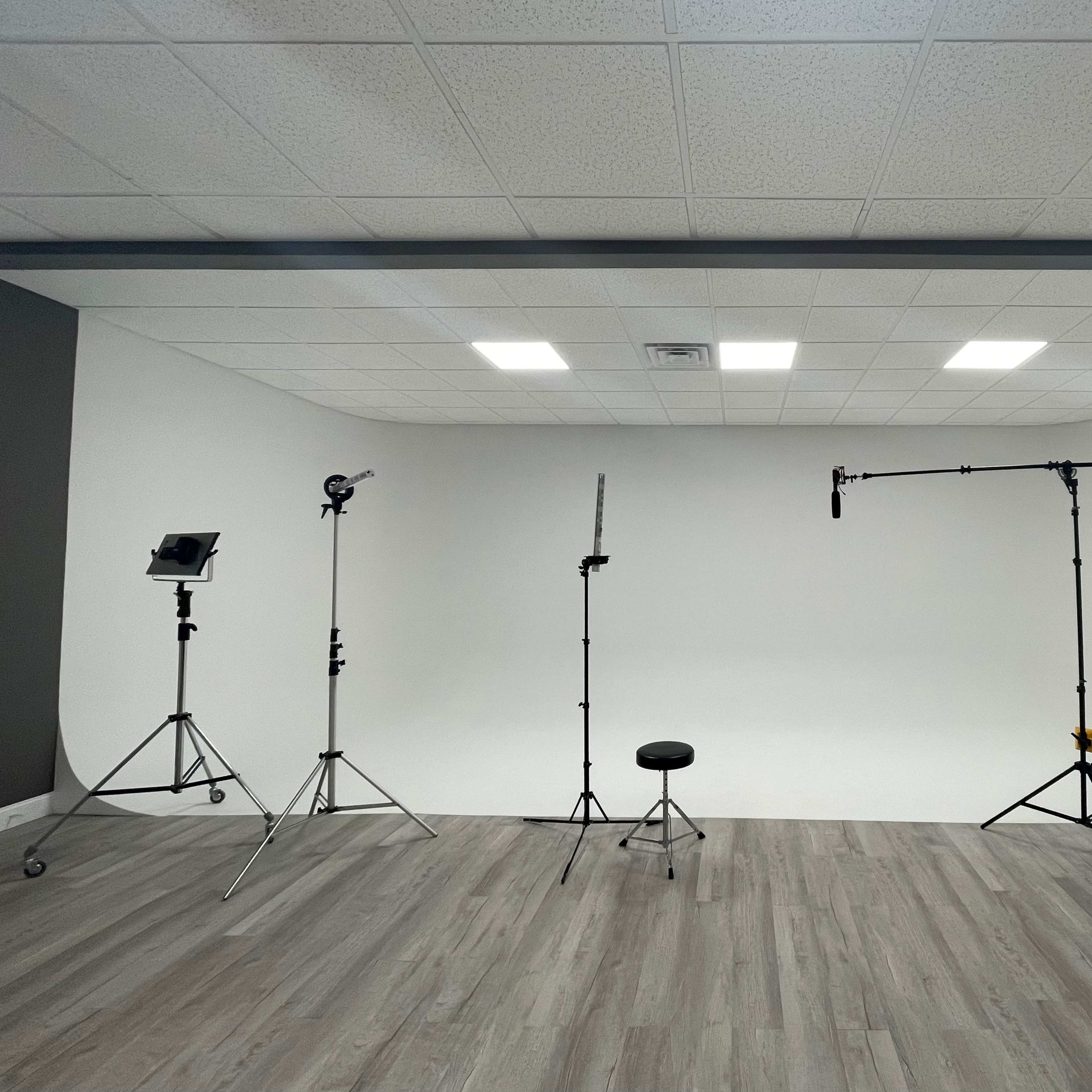 A photography studio with a white backdrop, various lighting equipment, and a single black stool arranged on a wooden floor.