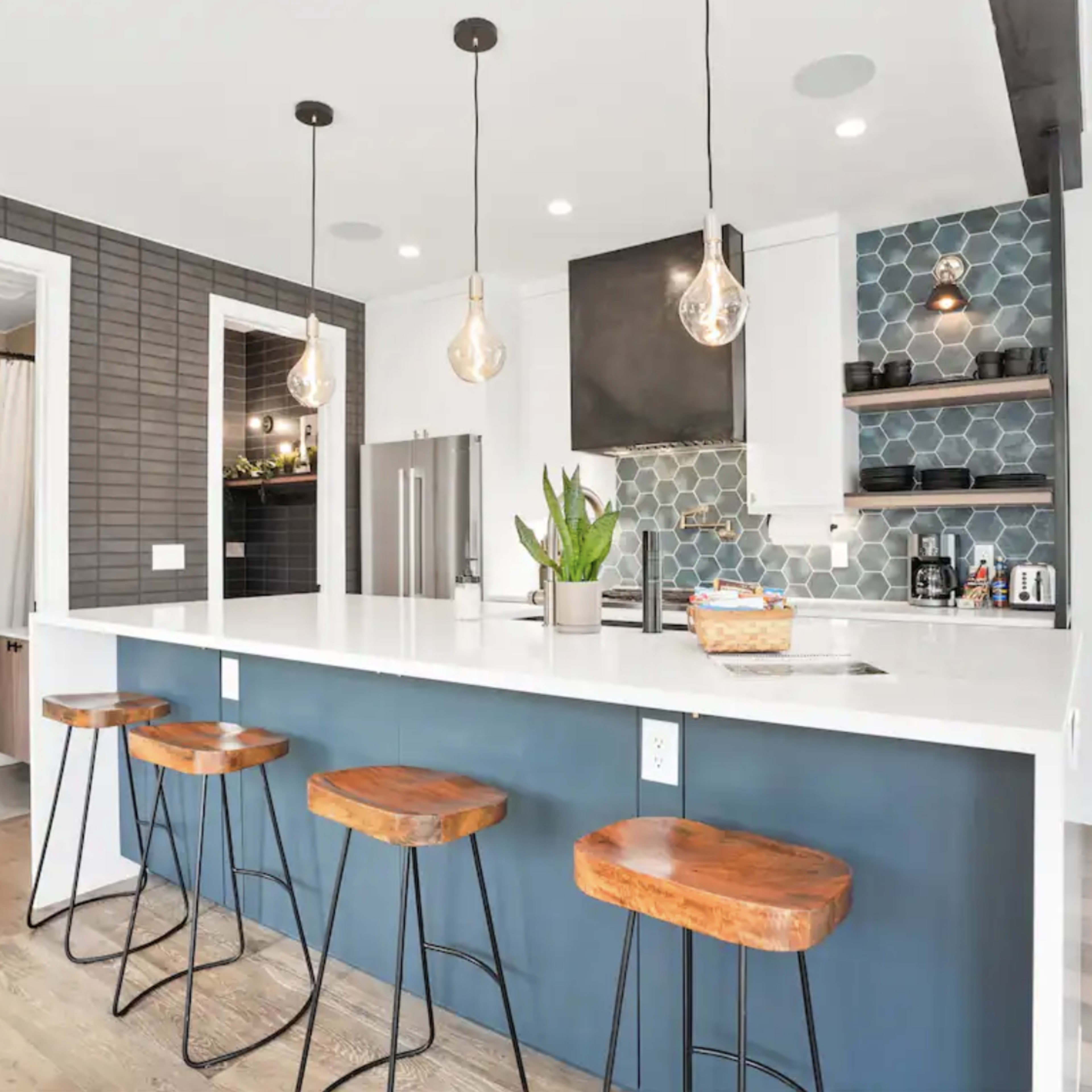A modern kitchen with a large white countertop, four wooden bar stools, and geometric tile accents on the walls.