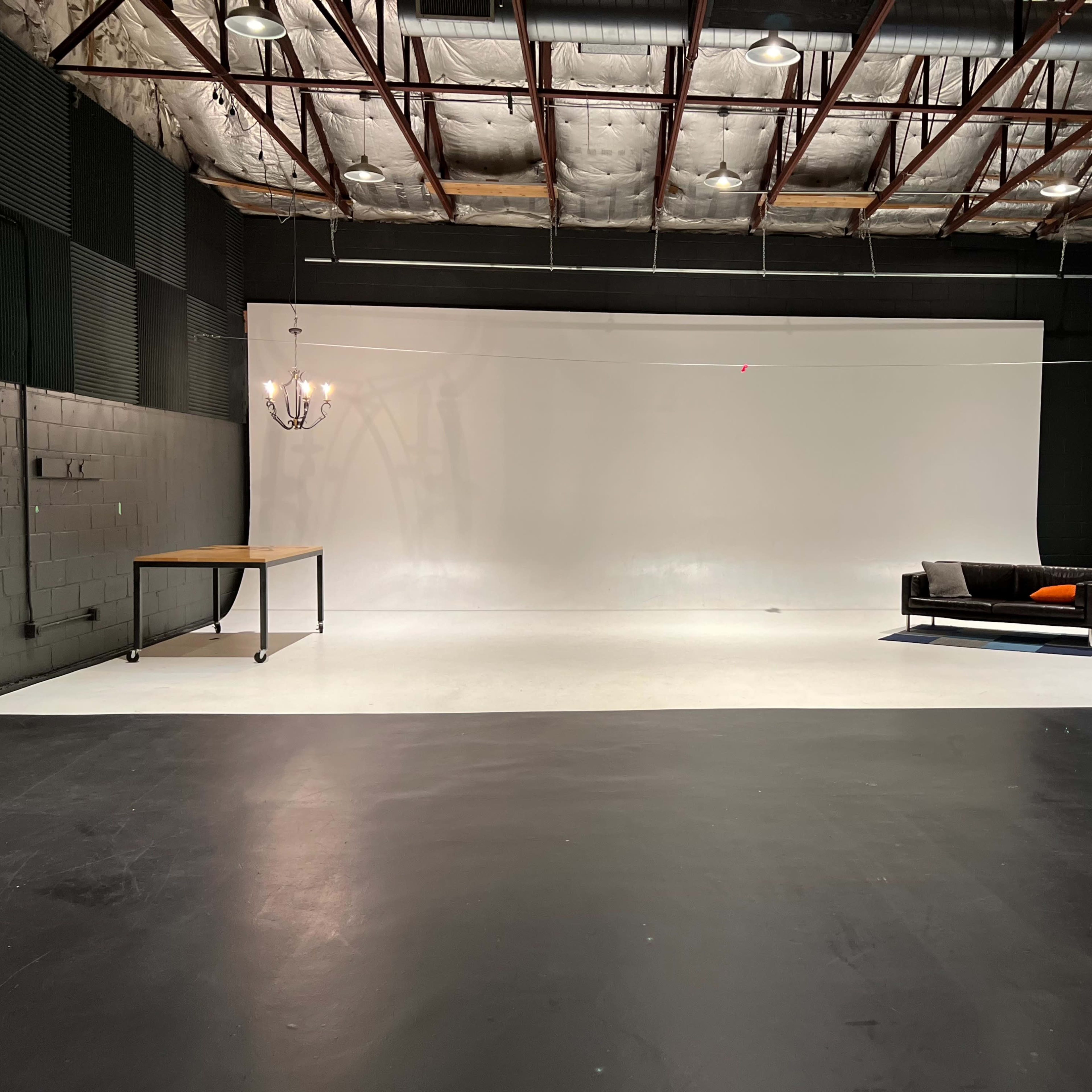 A spacious studio with a white backdrop, a wooden table, a black sofa, and a blue cabinet against the wall.