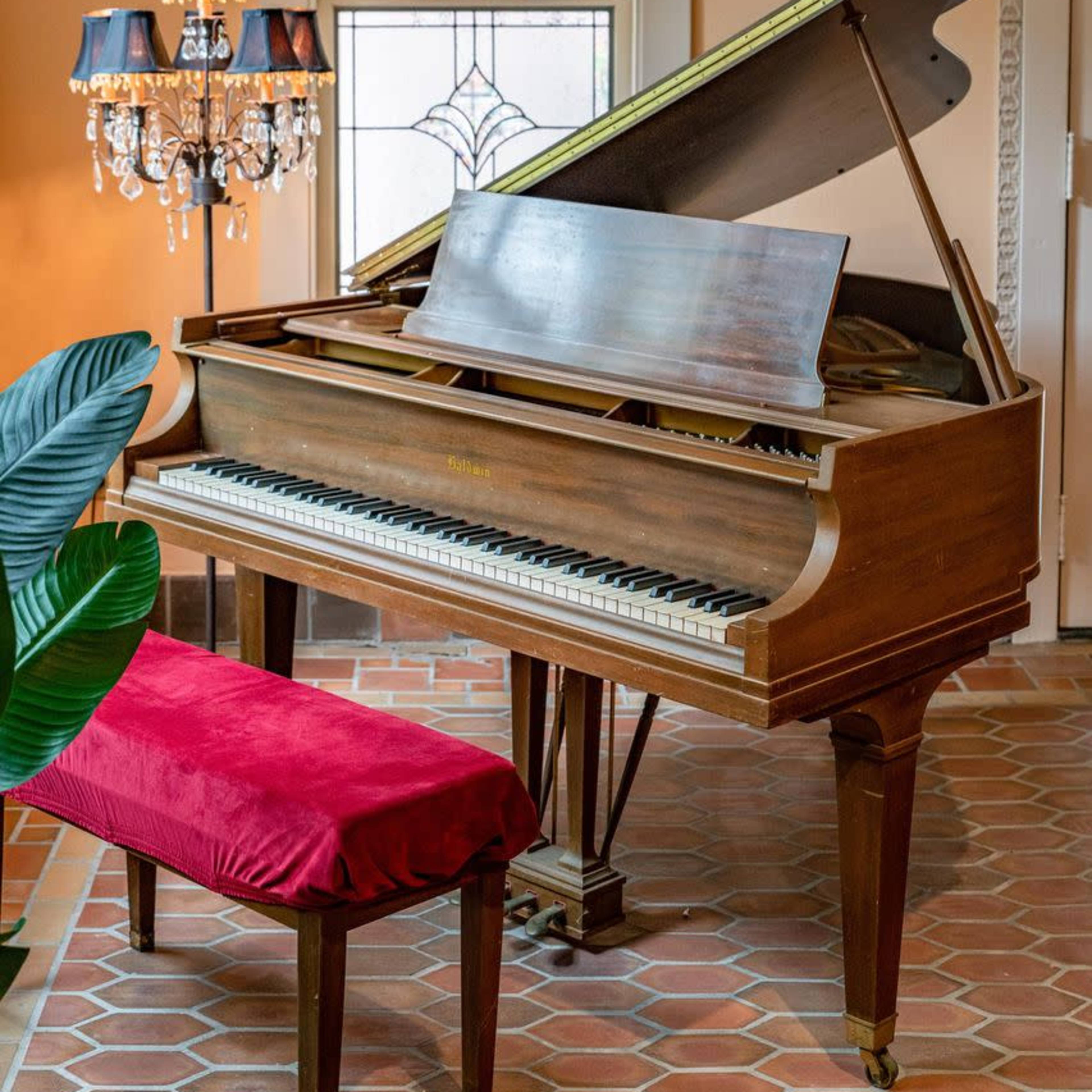 A grand piano with a raised lid sits beside a velvet-covered bench and a decorative lamp in a sunlit room.