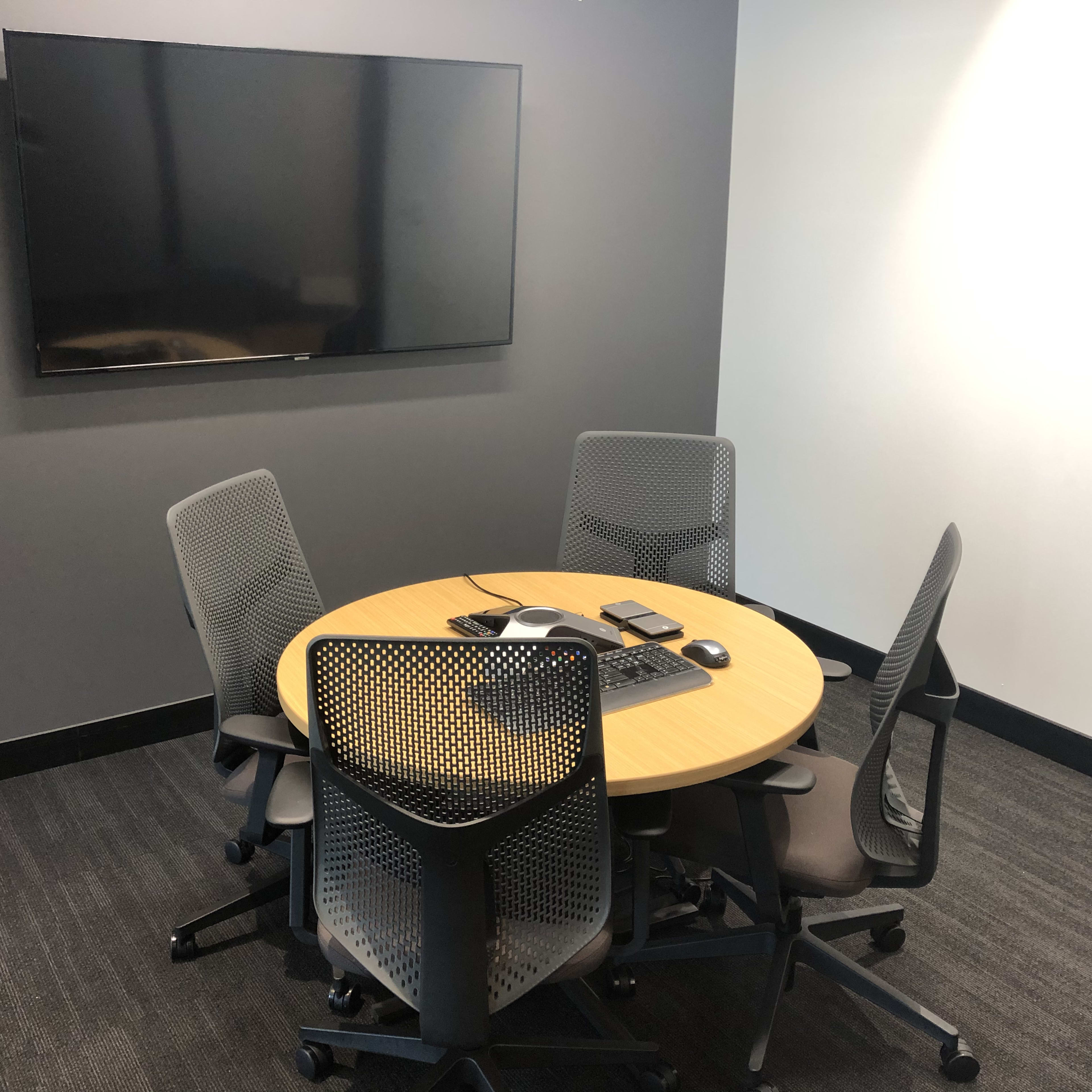 A small conference room features a round table surrounded by four mesh chairs and a wall-mounted television.