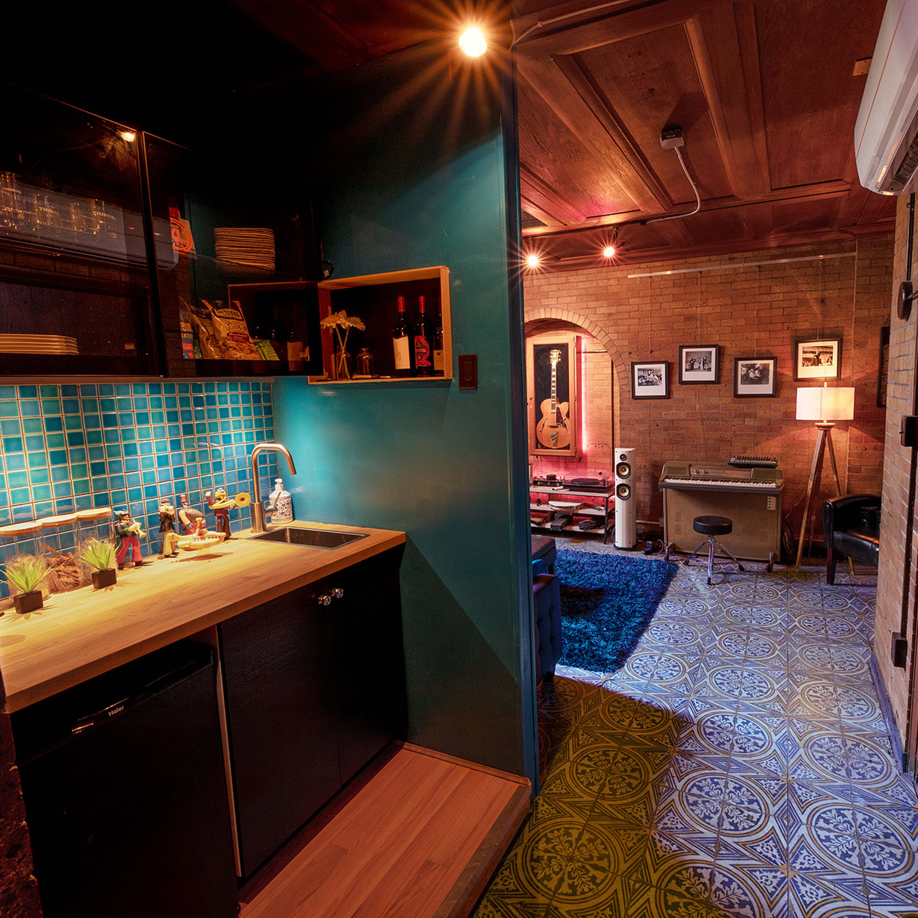 A cozy room with a small kitchenette on one side and a living area featuring a guitar, speakers, and decorative elements on the other side, all set against brick walls and patterned flooring.