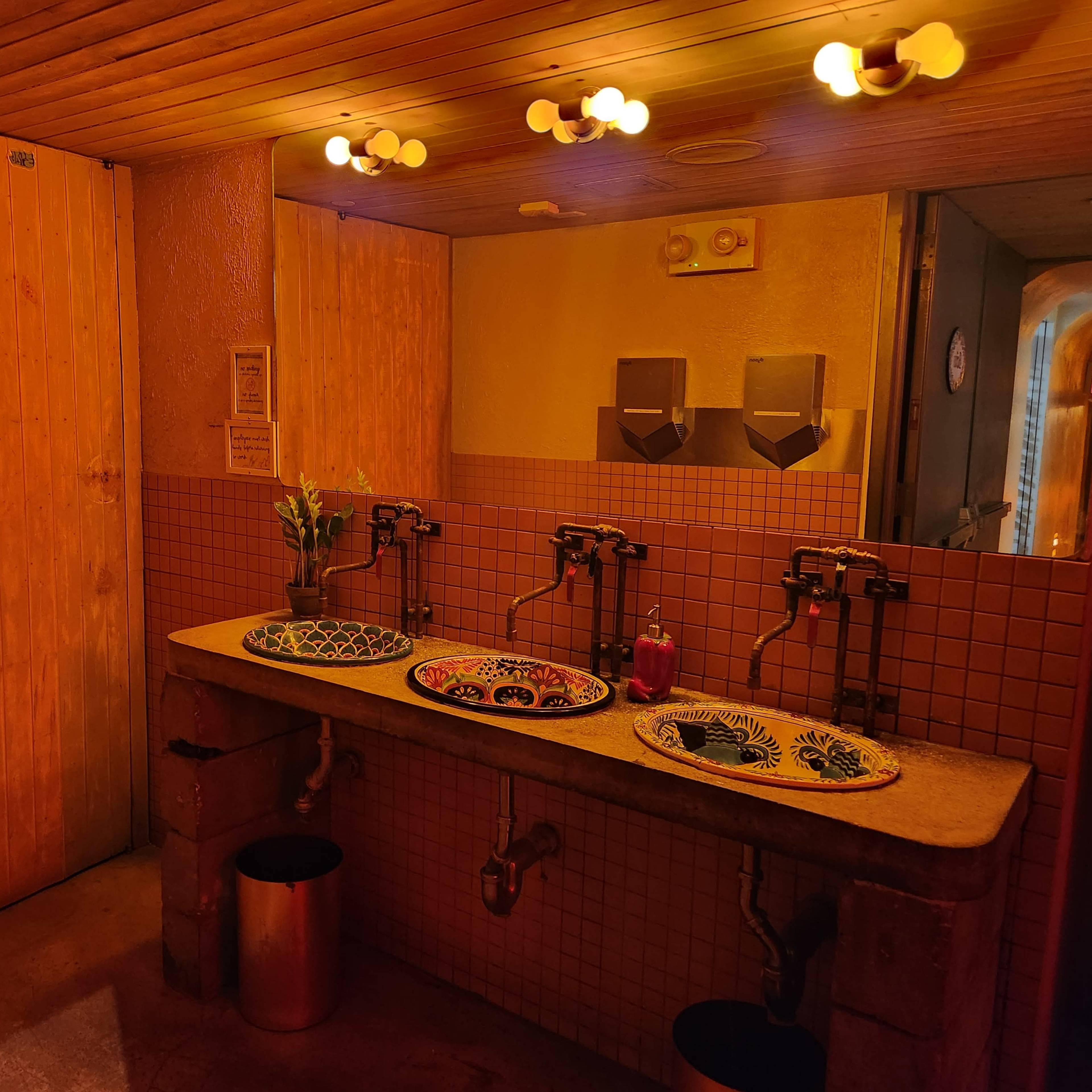 A restroom with three sinks, adorned with colorful bowls, and wooden-paneled walls illuminated by warm lights.