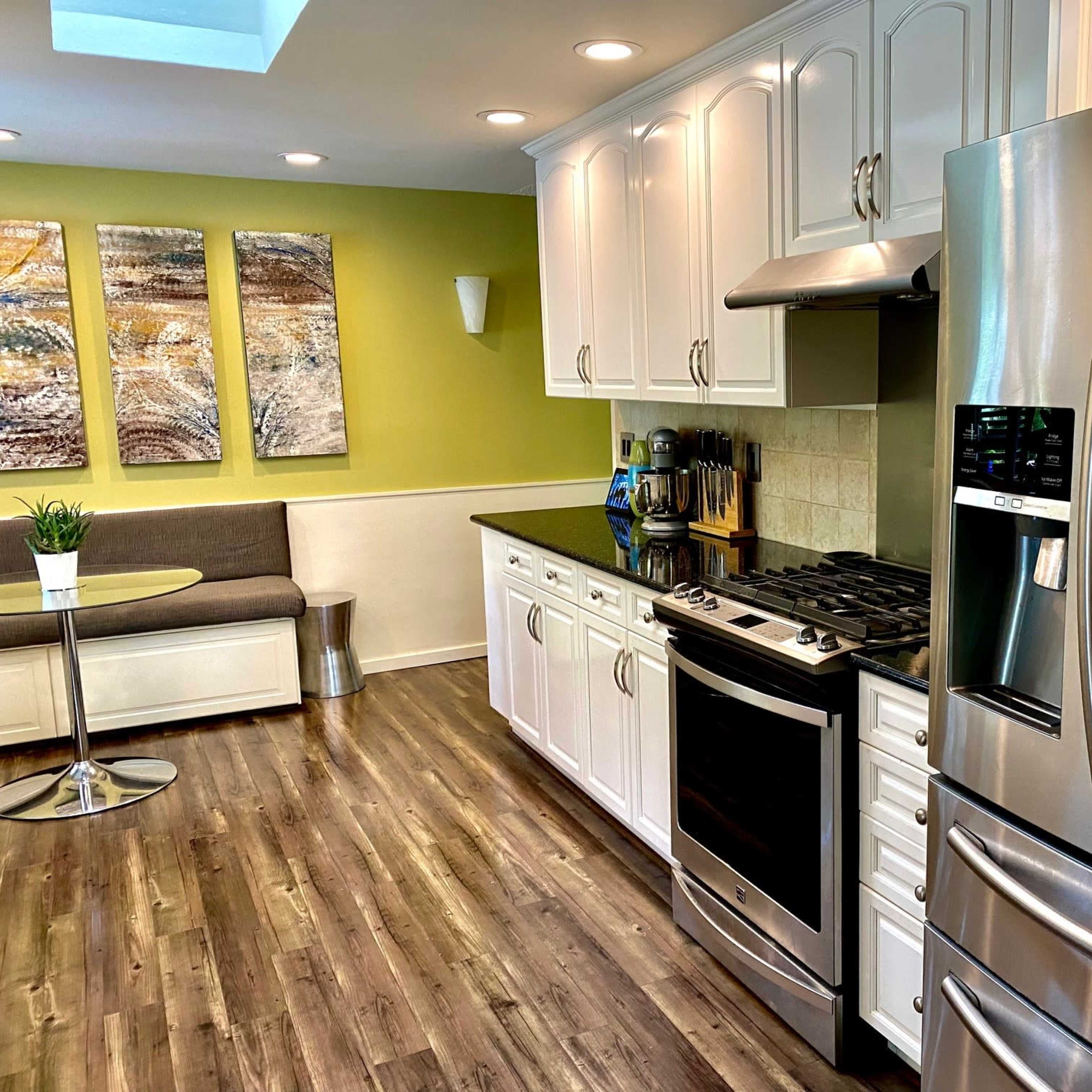 A modern kitchen with white cabinetry, a stainless steel refrigerator, a cooking range, and a small dining table in a corner.