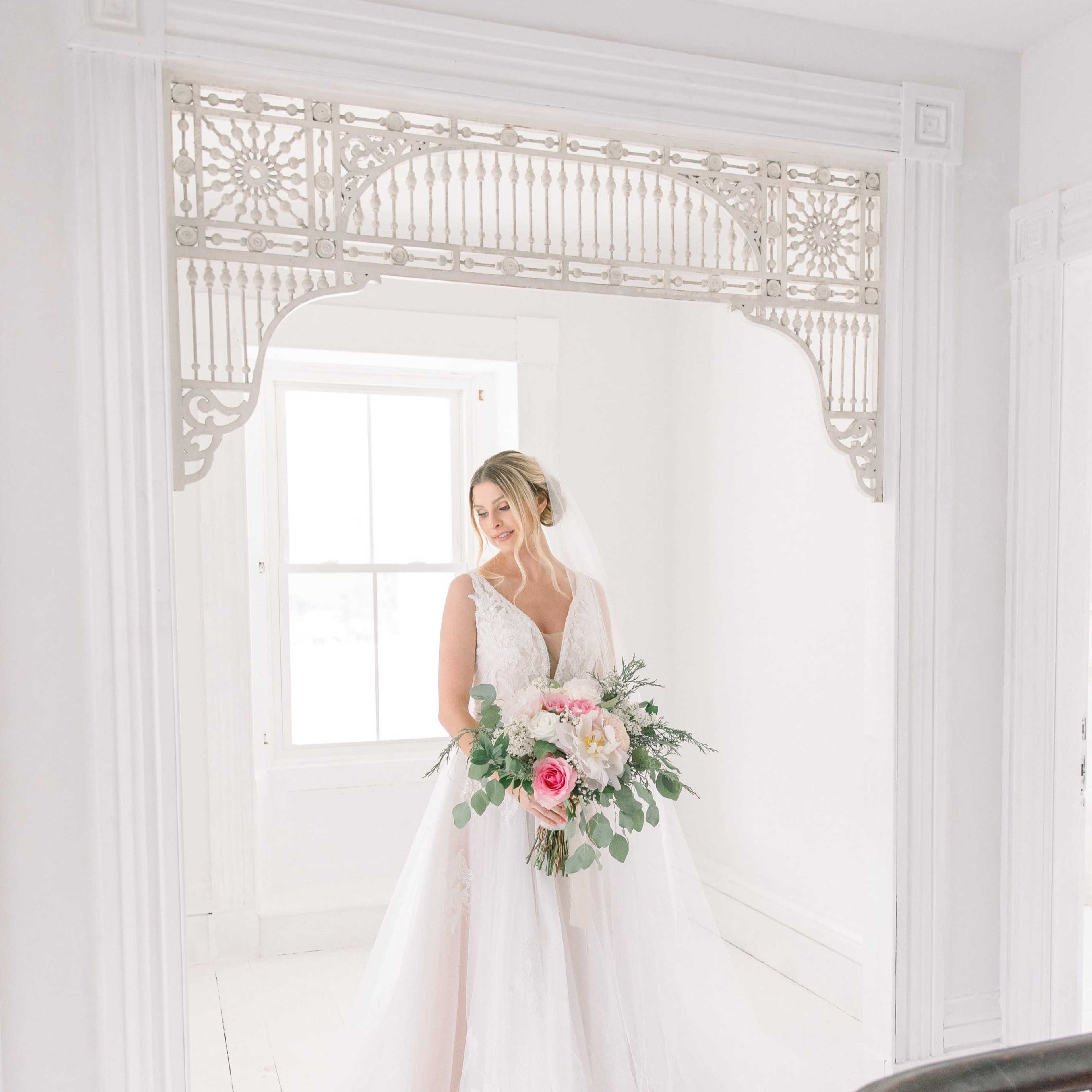 A bride holding a bouquet stands in a bright room adorned with ornate trim.