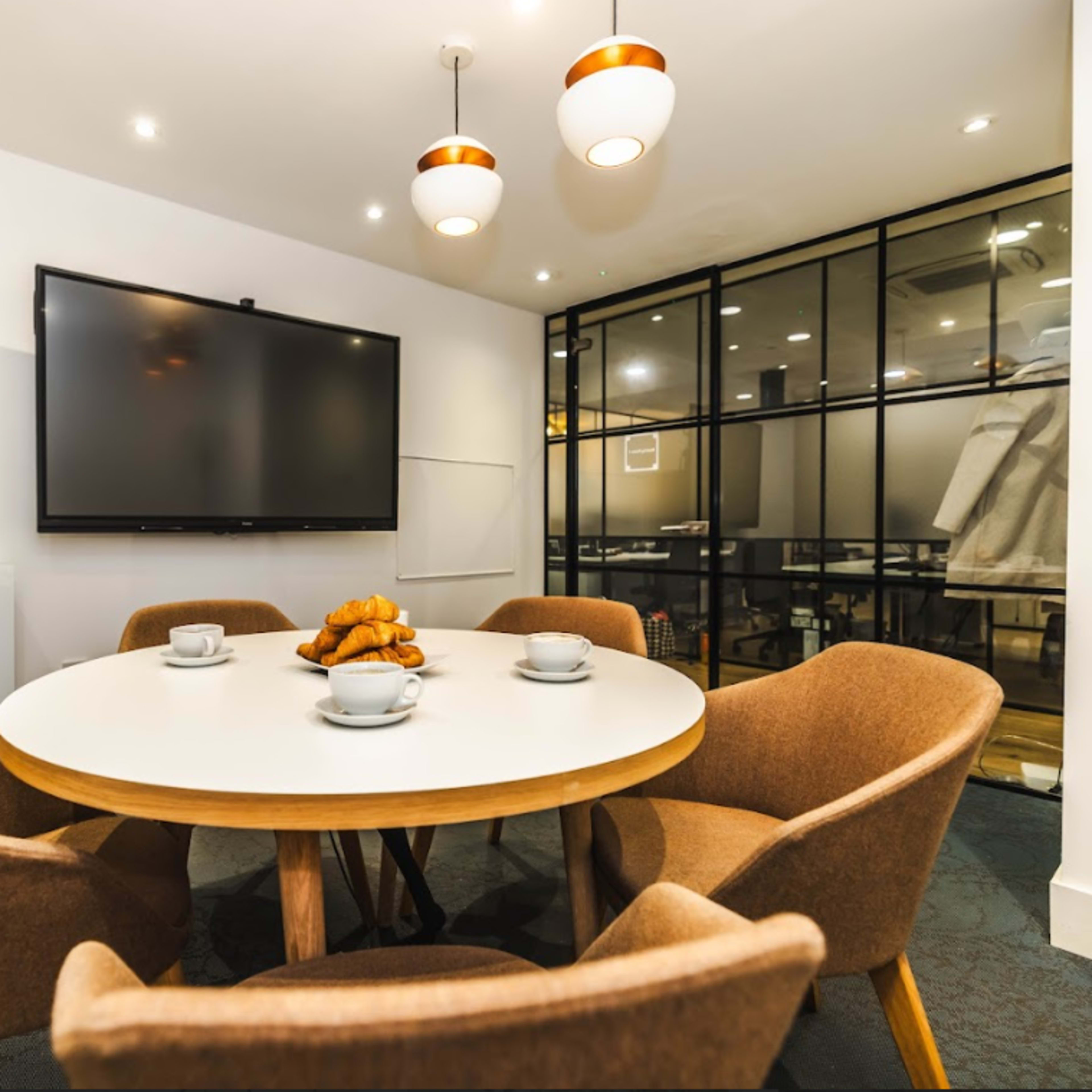 A round table with four upholstered chairs and a television mounted on the wall are arranged in a modern meeting room with a glass partition.