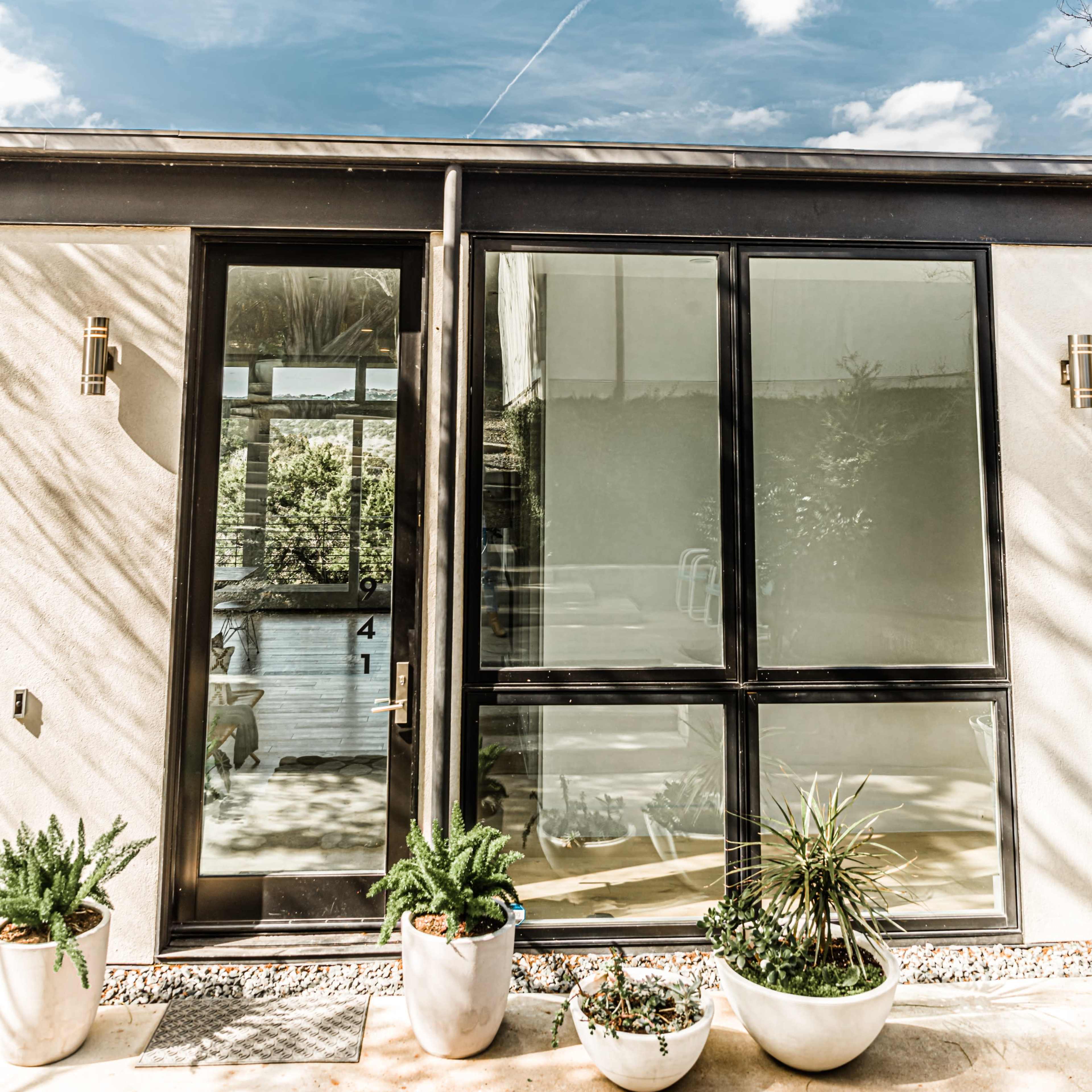 A modern house facade features large glass panels and potted plants under a blue sky with scattered clouds.