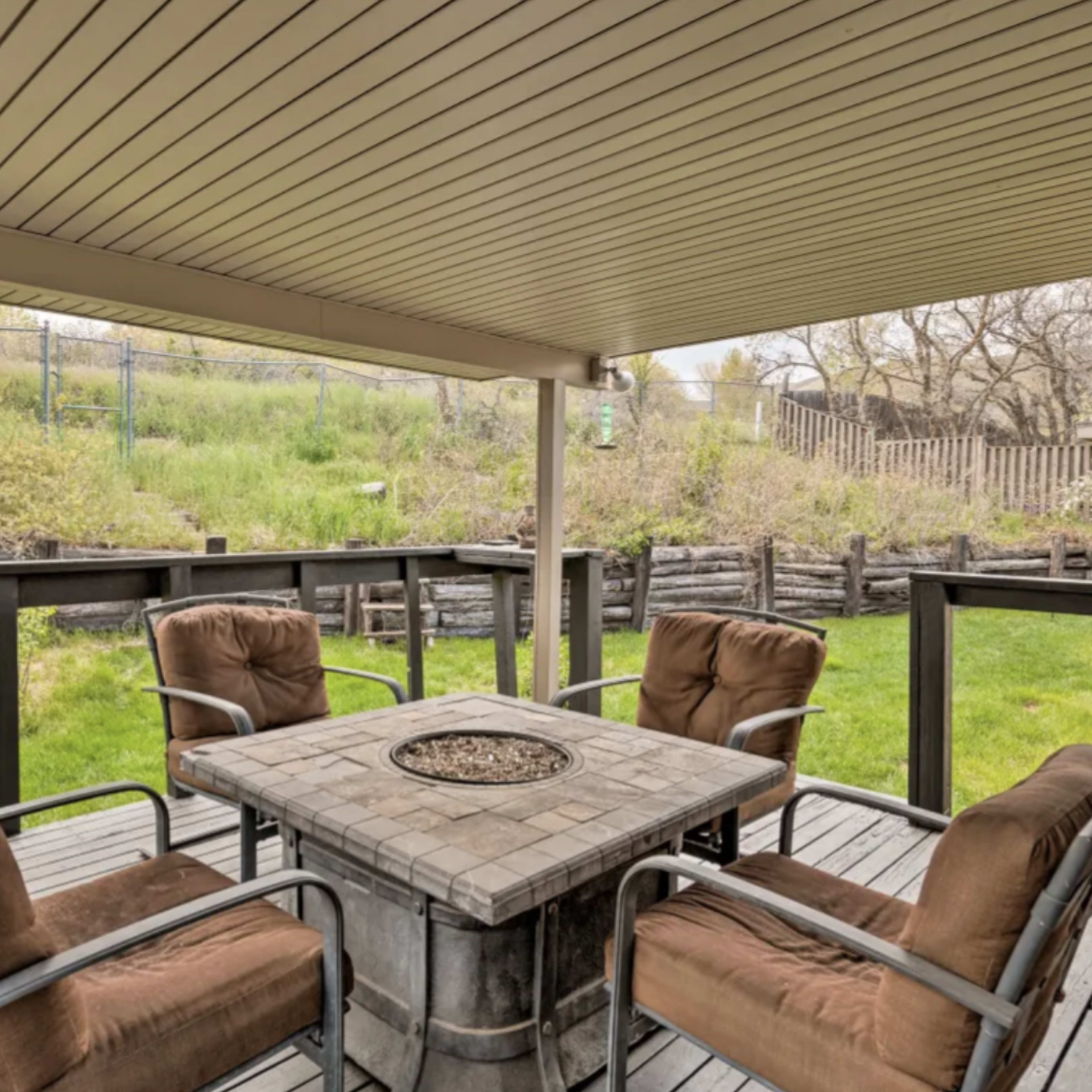 A patio area features a stone fire pit surrounded by four cushioned chairs and a view of a grassy landscape.