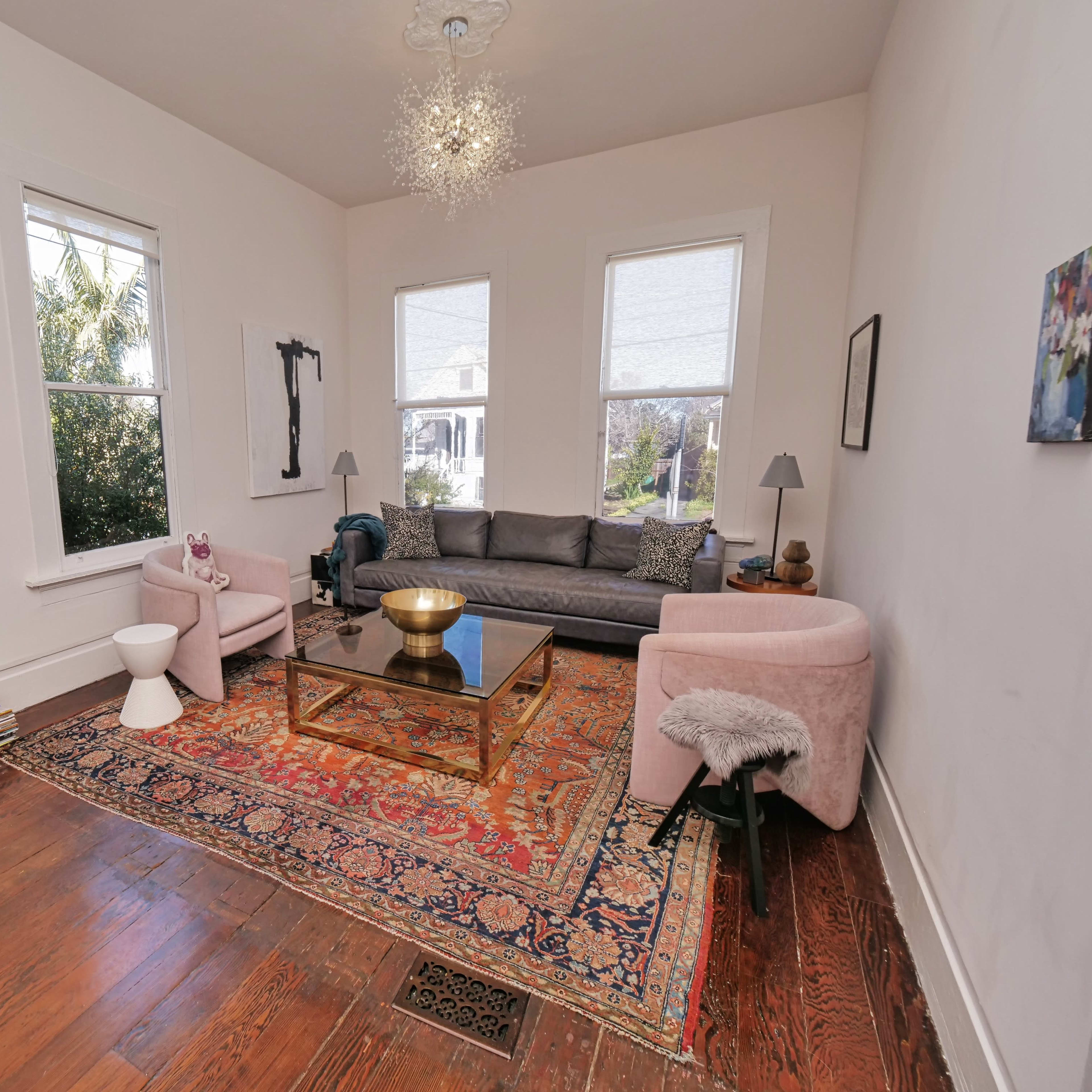 A living room with a gray sofa, two light pink chairs, a glass coffee table, and a tall stack of books next to a window.