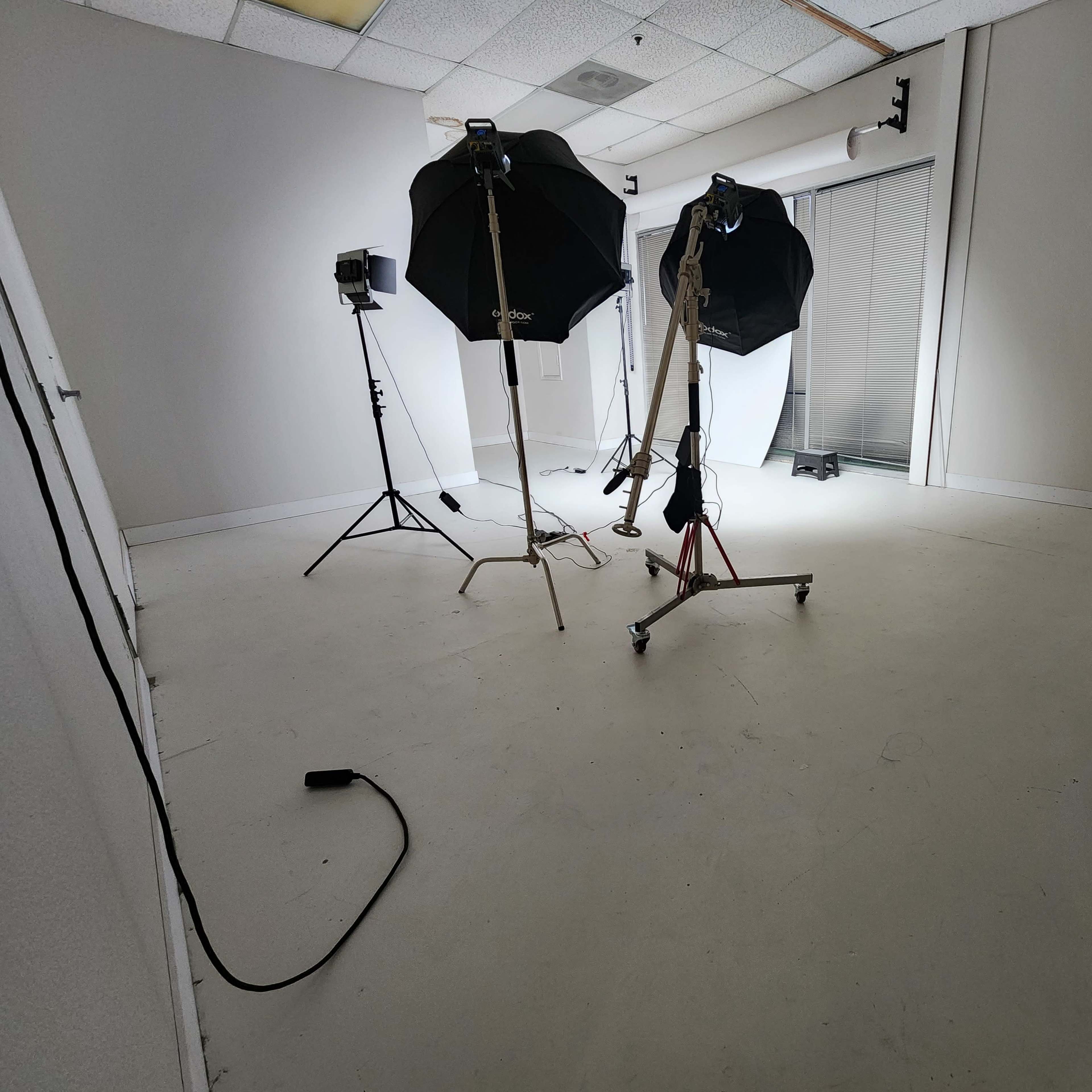 A sparsely furnished studio space with two softbox light stands and a tripod, set against white walls and a large window.