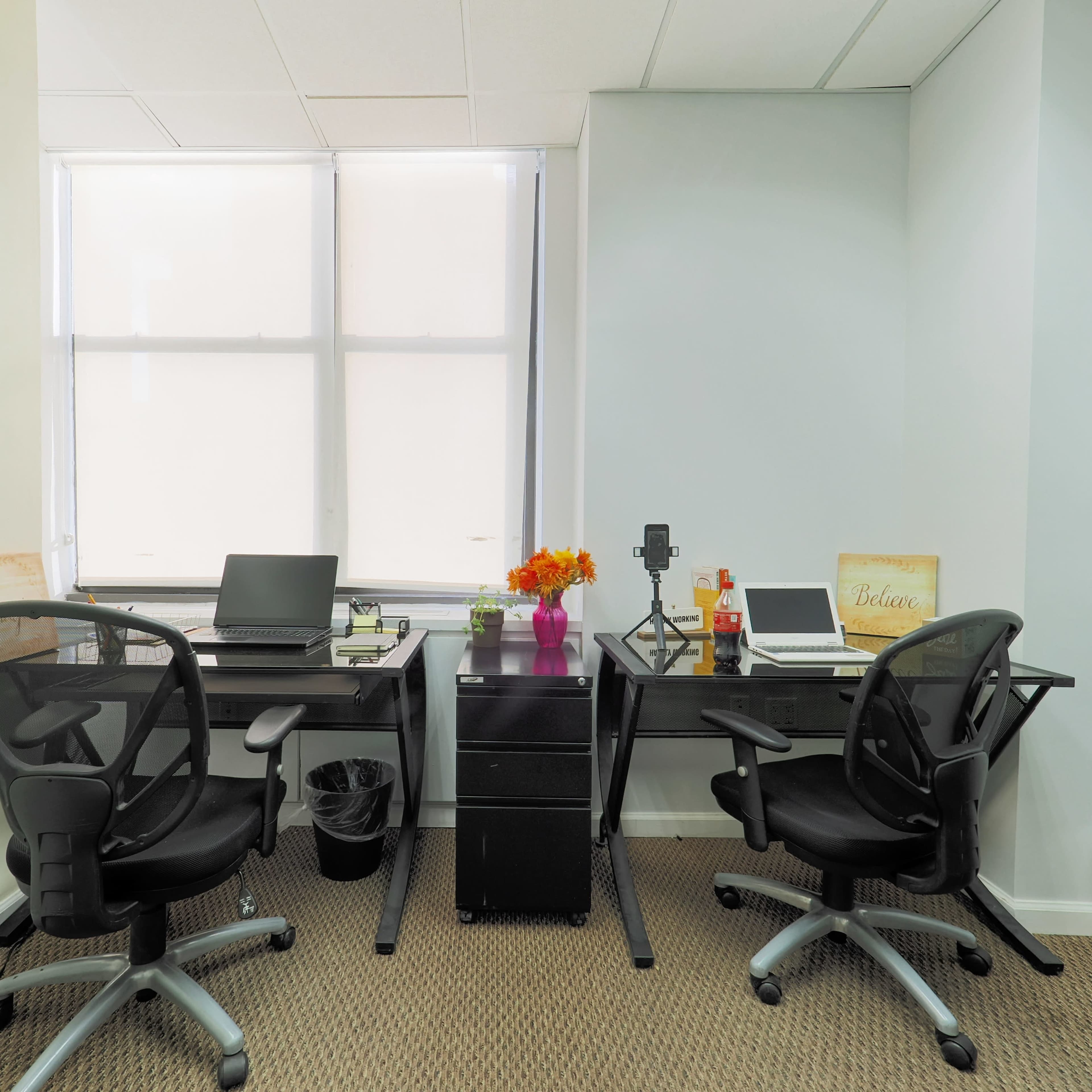 A small office space features two desks with laptops, a flower vase, and a filing cabinet, along with ergonomic chairs and a window with blinds.